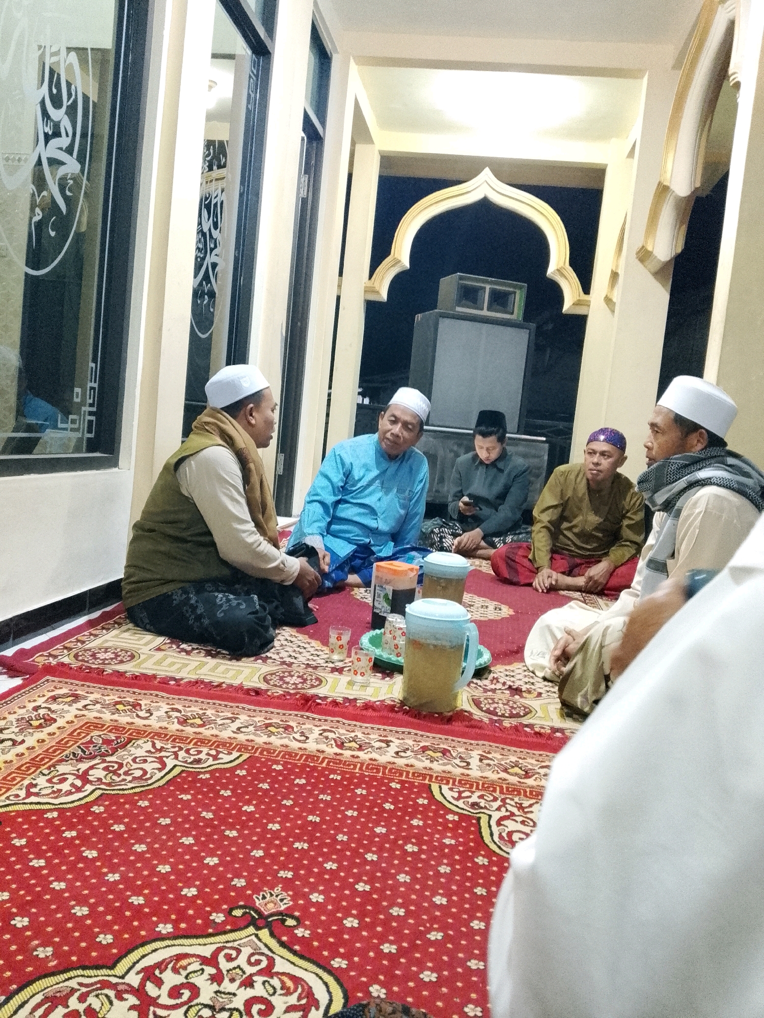 In a room with a red and gold patterned carpet, six men are seated on the floor. Three men are wearing white skullcaps, and one is wearing a purple skullcap. One man in a green vest and brown scarf is in the foreground, to the left, facing right. Another man in a blue shirt and white skullcap is to his left, facing him. The other three men are seated further back. One man in a black shirt and white skullcap is looking down at a phone. Another man in a brown shirt and striped scarf is facing left. A sixth man, only partially visible, is in the right foreground, wearing a white garment and a white skullcap. In front of the men, there are two pitchers of drinks, a plate with small glasses, and a small green dish. The room has architectural elements such as arched doorways and columns, and there is calligraphy on the windows. The overall atmosphere suggests a gathering or meeting.