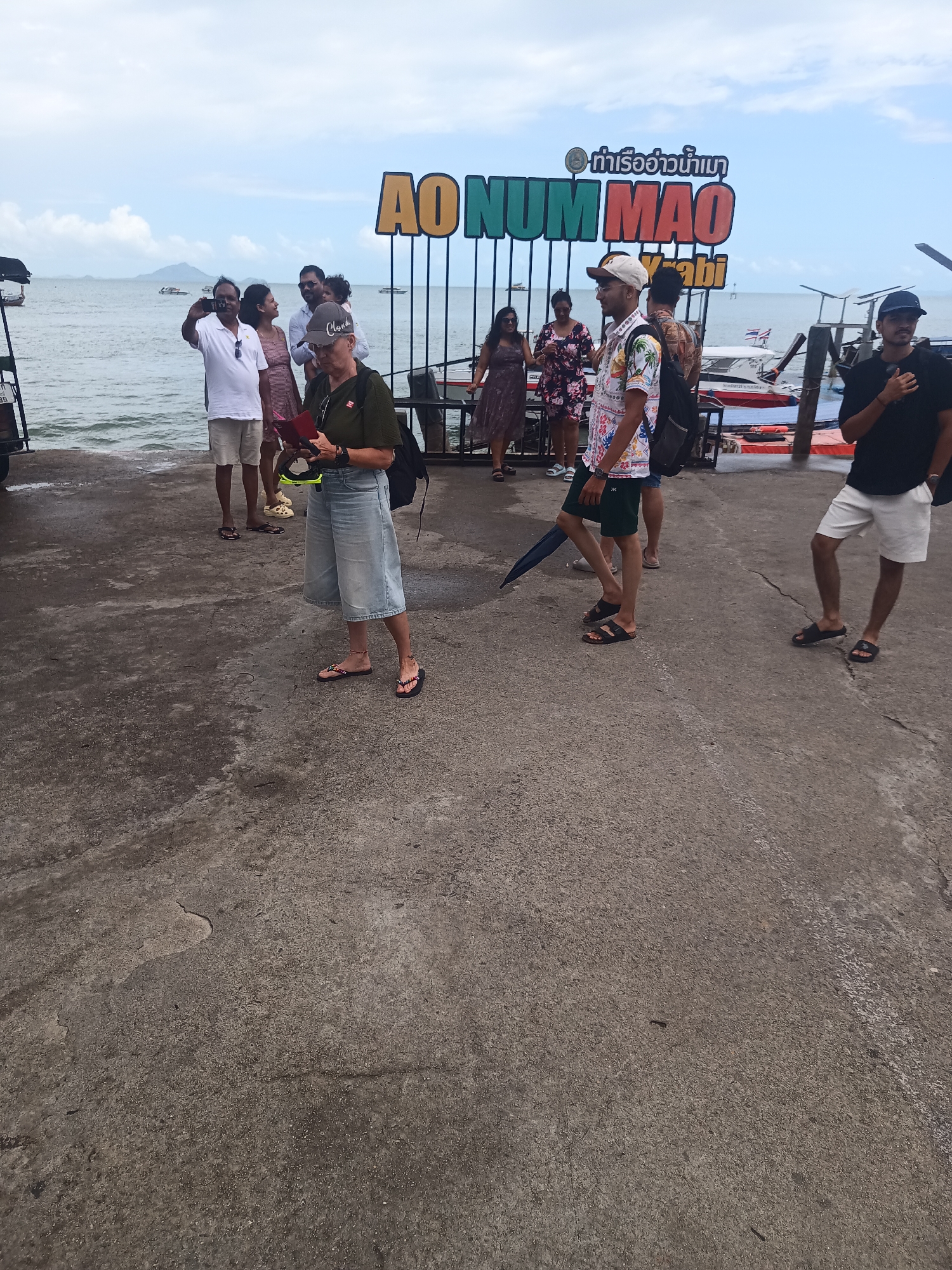 The image depicts a group of people gathered at a pier in Ao Num Mao, Thailand, as indicated by the sign in the background. The sign is predominantly green and red and also features Thai script. Several boats are docked nearby. The group consists of approximately ten individuals. They are dressed in casual attire, including shorts, shirts, dresses, and sandals. Some are interacting with each other, while others appear to be looking out at the water. A woman in the foreground is wearing a green shirt and jean culottes; she is reading a pamphlet. The pier itself is made of concrete and has a weathered appearance. The weather appears overcast.