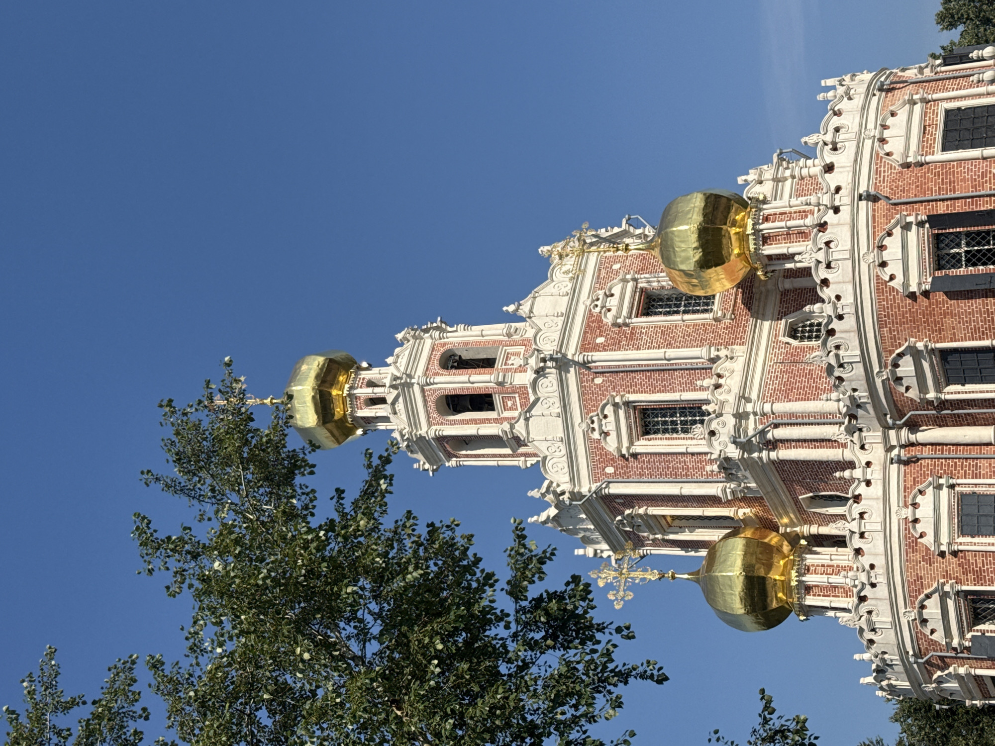 The image shows a close-up, low-angle view of the Novodevichy Convent in Moscow, Russia, under a clear blue sky. The bell tower and several golden domes of the convent are prominently featured, partially obscured by the leaves and branches of a tree in the foreground. The architecture is characterized by red brickwork, white decorative elements, and ornate golden cupolas topped with crosses. The sun illuminates the structure, highlighting its intricate details and the reflective surfaces of the domes. No people are visible in the frame.