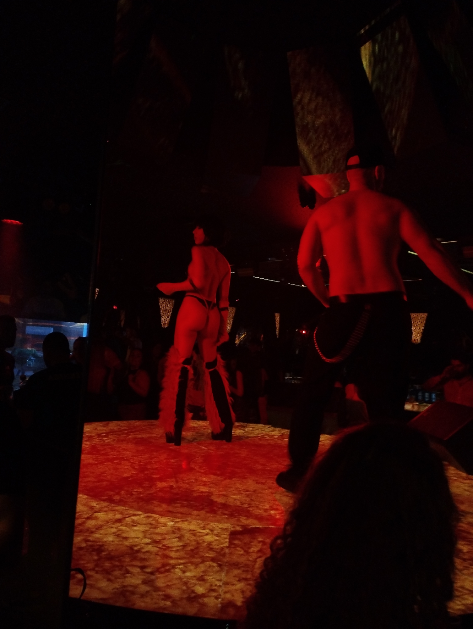 A stage at a dimly lit venue in Unknown, Unknown, shows a woman and a man, both facing away from the camera. The woman, wearing a thong and what appears to be furry leg warmers, stands on the left side of the stage. To the right is a shirtless man with a black cap, wearing black pants with a chain detail. A crowd is visible in the background, and red lighting dominates the scene.