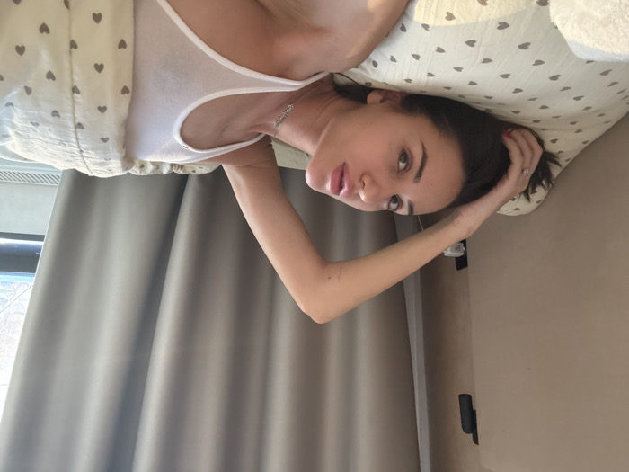 A woman in a white tank top lies on a heart-patterned pillow, her dark hair tousled and one hand touching her head. She gazes at the camera with a slight smile. A beige curtain hangs in the background near a window. The wall on the right is a light beige color.