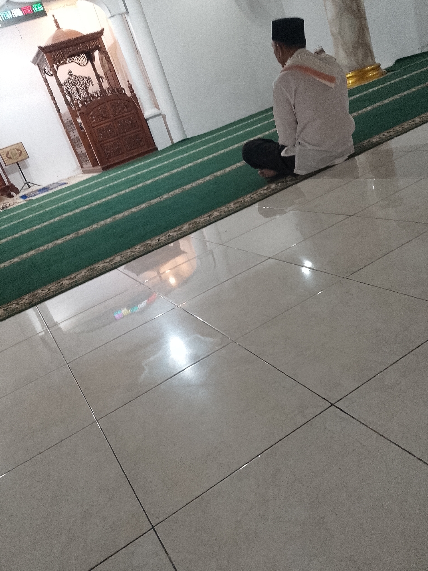 In the city of Ranoyapo, Indonesia, within a mosque, a man is seated on a green carpet, facing away from the camera. He is wearing a white long-sleeved shirt, black pants, and a black cap. A white scarf with orange trim is draped over his shoulders. The mosque features a traditional wooden pulpit on the left side, carved with intricate designs. The floor is tiled in a light color and reflects the ambient light. The scene suggests a moment of prayer or contemplation.