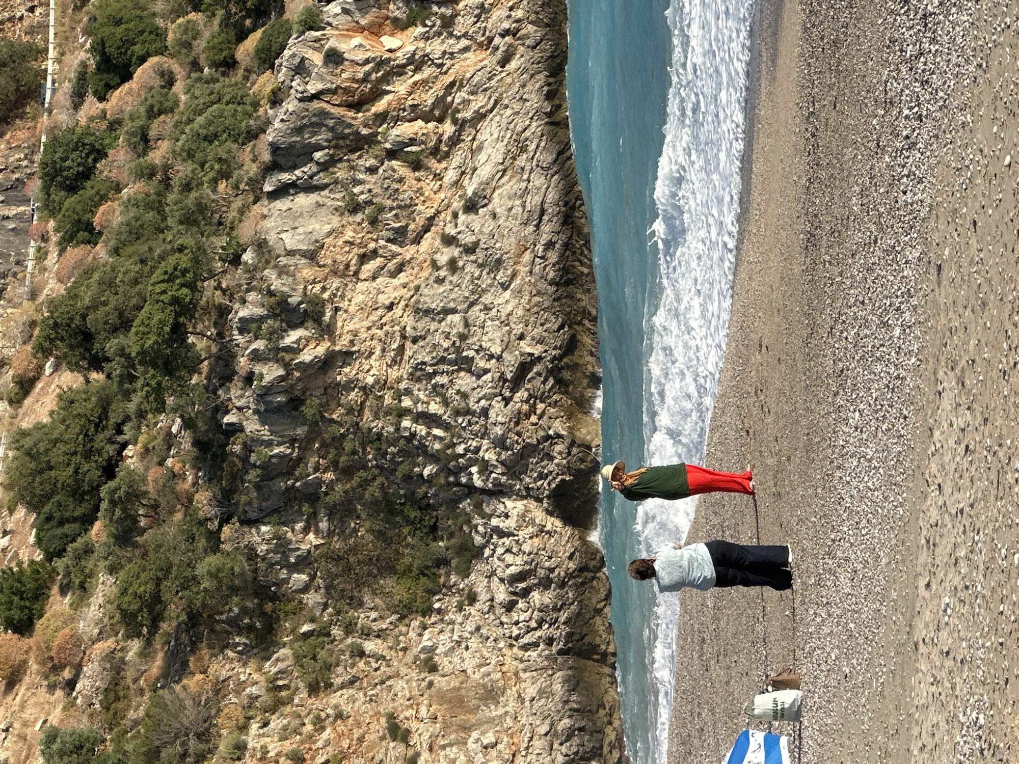 This photograph captures a scene on a beach in Fethiye, Turkey. The beach is pebble-covered and stretches to meet the turquoise water of the sea, which is fringed with white waves. The beach is backed by a steep, rocky cliff covered with vegetation. Two individuals are present on the beach. One person is wearing a hat, a green top, and red trousers. The second person is wearing a light-colored top and dark trousers. A bag and a blue and white object are near the second person. The overall scene conveys a relaxed, coastal environment.