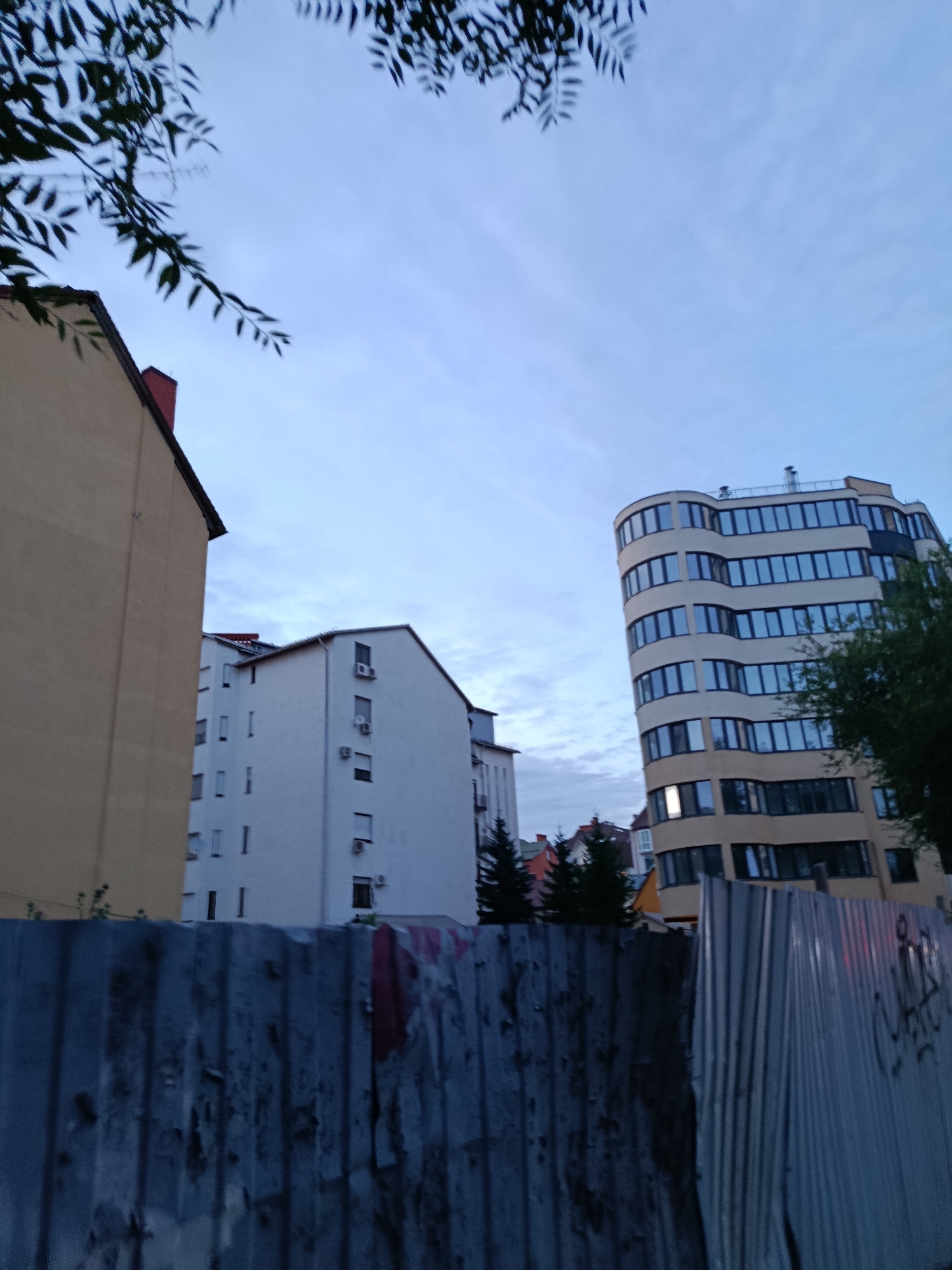 This image depicts an urban scene, likely taken during dusk, with a focus on buildings and a corrugated metal fence in the foreground. On the left, a tall, cream-colored building with a red-tiled roof and a prominent chimney is partially visible. Adjacent to it, to the right, is a white building with multiple windows and air conditioning units mounted on its facade. Further to the right, a modern, curved building with a light-colored exterior and numerous dark-framed windows dominates the upper right portion of the image. A lush green tree partially obscures this building. In the background, several smaller buildings with red roofs are visible, along with evergreen trees. The sky is overcast with scattered clouds. The foreground is characterized by a weathered, grey corrugated metal fence, which appears to be a construction site barrier, with some graffiti visible on its surface. The overall impression is a view of urban architecture, possibly in a developing area, given the presence of the fence. The precise location in Saratov, Russia, cannot be determined from the visual information alone.