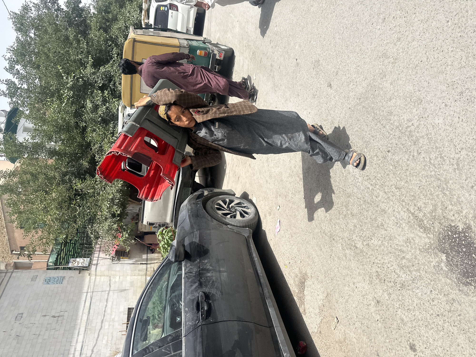 A man, dressed in traditional clothing, is carrying a red object on his shoulder. He stands on a paved street with a black car beside him. In the background, another man in maroon clothing stands near a green and yellow vehicle. The location is Quetta City Tehsil, Pakistan. A tree and buildings are visible in the background.