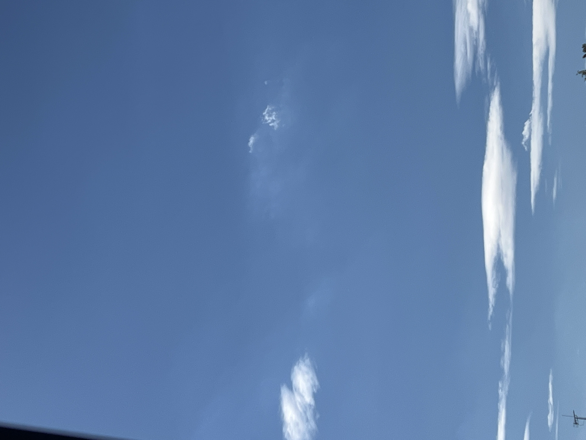 The image shows a clear blue sky with scattered white clouds. In the foreground, a tall antenna-like structure is partially visible on the left side, suggesting an outdoor urban environment. The clouds are wispy and thin, with larger formations concentrated towards the bottom of the frame. The overall impression is of a bright, sunny day. There are no people or specific landmarks visible to pinpoint the location to Warsaw, Poland.