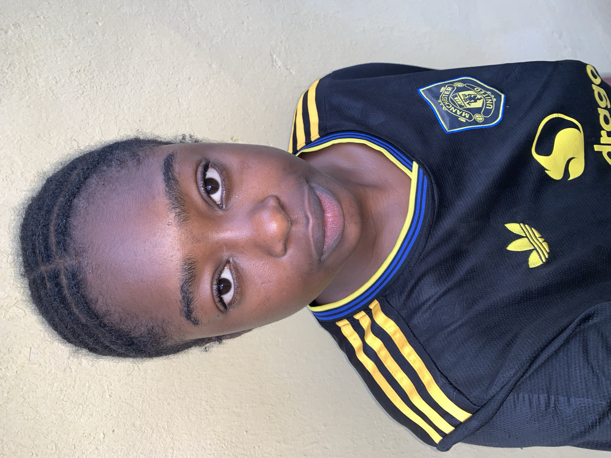 A medium indoor shot depicts a person with dark skin and black hair styled in braids, wearing a black Adidas soccer jersey with yellow stripes and trim. The jersey features a "Manchester United" emblem on the upper left and "dragon" branding on the right. The person is positioned in front of a plain, light beige wall. The location is not specified, but the prompt includes the city of Rafin Par, Nigeria.