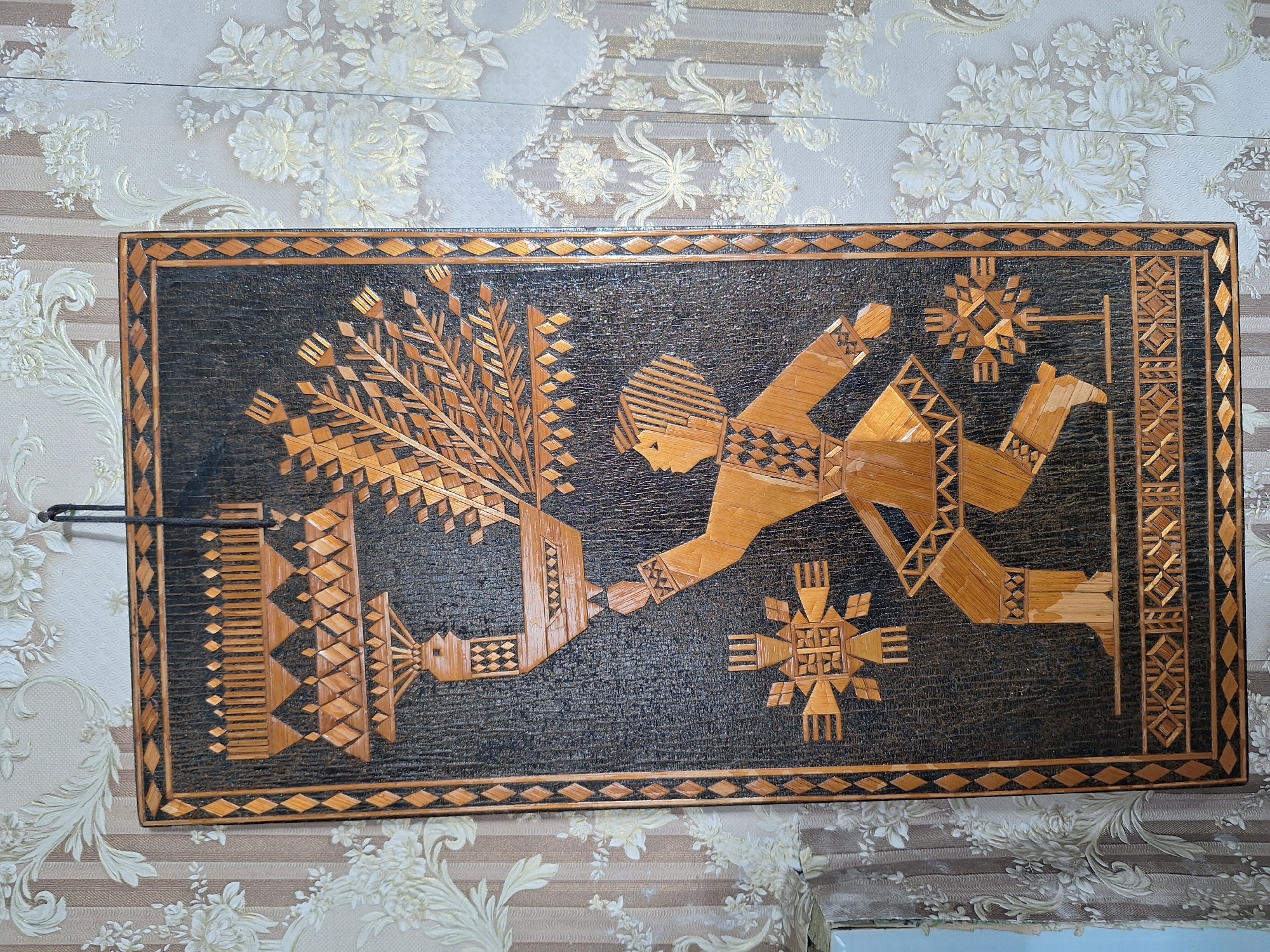 The image shows a straw-crafted wall hanging featuring a stylized human figure carrying a large bird with an elaborate headdress and fanned tail. The figure is depicted in a dynamic pose, seemingly walking or dancing, adorned in clothing with geometric patterns. Two star-like ornaments are positioned on either side of the figure. The artwork is framed with a border of woven straw and mounted against a backdrop of patterned wallpaper. The wall hanging is suspended by a dark cord. The overall scene is an interior setting.