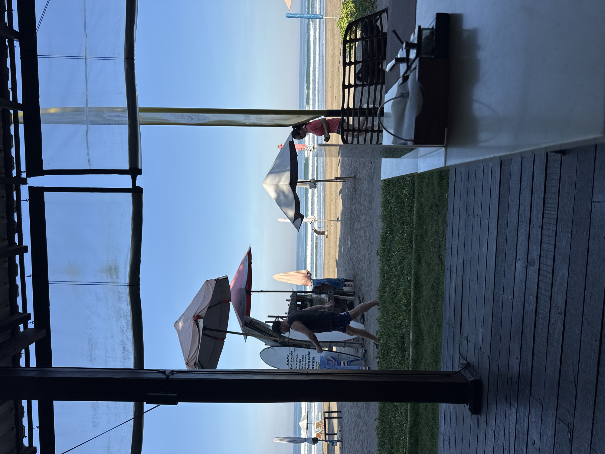 The image depicts a view of the beach in Kuta, Indonesia, as seen from a partially covered patio or deck area. The deck is made of dark wood planks. Outside the deck area is a sandy beach with several umbrellas, surfboards and people. A man in dark clothes appears to be demonstrating a surfing move. The ocean is visible in the background, with gentle waves. The sky is clear and blue. A clear plastic cover appears to be used as a window enclosure on the deck.