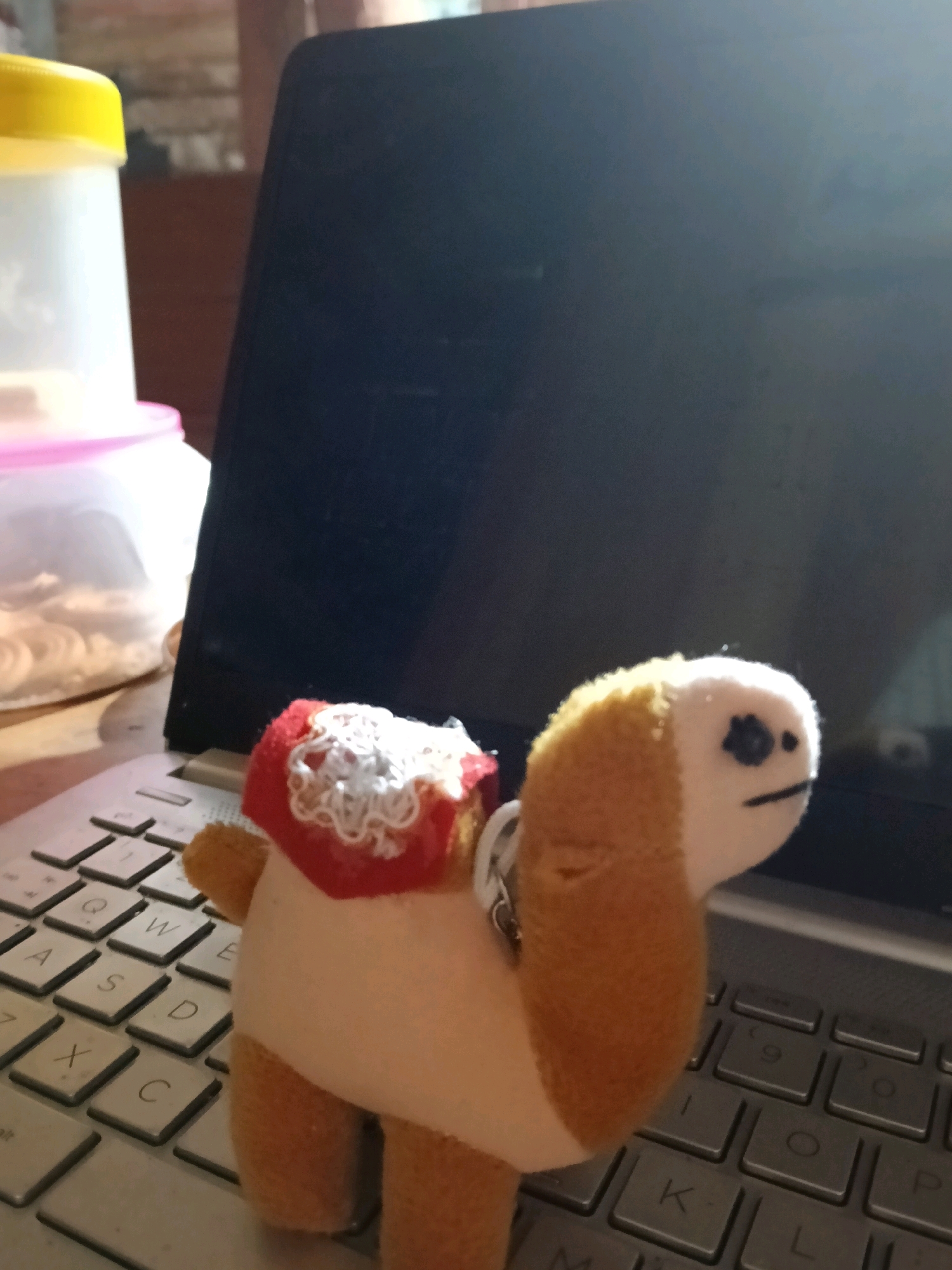 A stuffed camel toy with a red saddle decorated with white lace sits on a laptop keyboard. To the left of the laptop, a clear plastic container with a pink lid is visible, filled with what appears to be round, light-colored food items. Another white plastic container with a yellow lid is partially visible behind the first. The background suggests an indoor environment with some wooden furniture. The scene is likely a desk in an urban setting within Galuk, Indonesia.