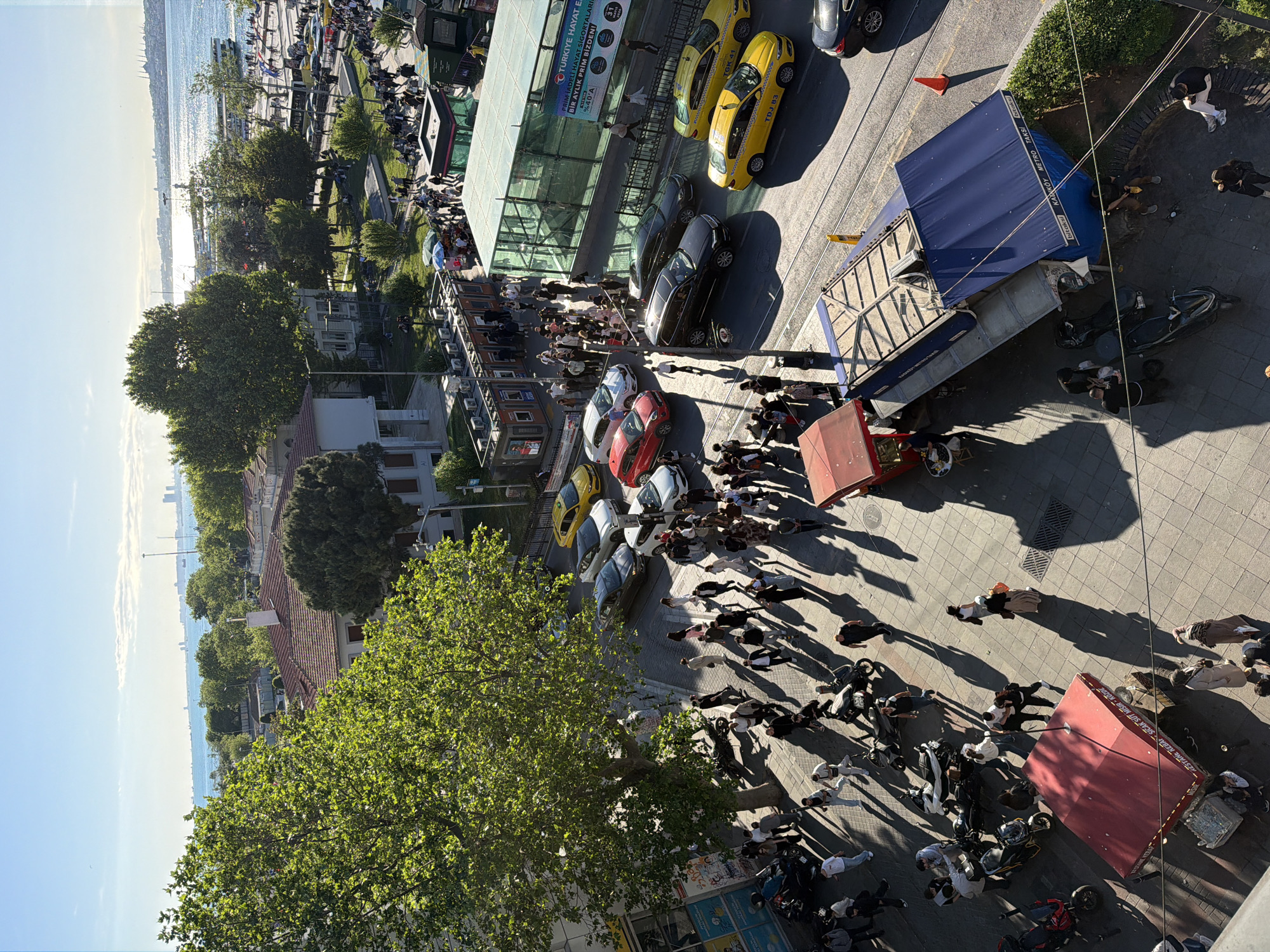 The image presents a bird's-eye view of a bustling street scene in Kadıköy, Turkey. The Marmara Sea is visible in the background. Several parked cars, including yellow taxis and other vehicles, line one side of the street. A large crowd of people is gathered in the center of the street, casting long shadows, suggesting a sunny day. Several individuals are moving within the crowd, with many wearing darker clothing. Two stalls with red awnings and a kiosk with a blue awning are present, and there is an entrance to the Kadikoy Metro. The street is paved with rectangular tiles. The surrounding area includes buildings, trees, and greenery.