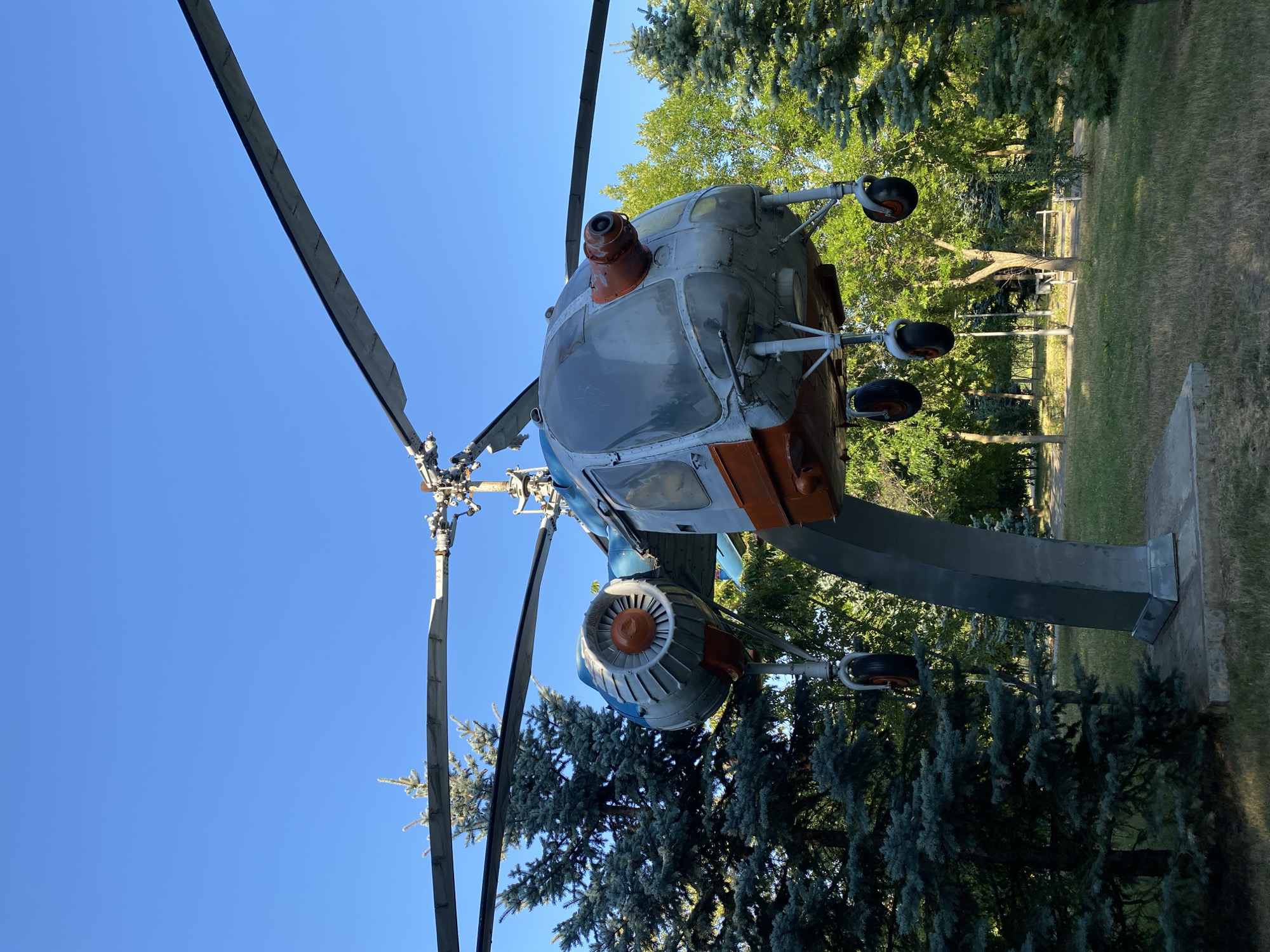 The image shows a parked helicopter outdoors, possibly as a monument or exhibit, against a clear blue sky. The helicopter is silver with orange accents and has a prominent rotor system. It is mounted on a large, curved, grey metal stand. The helicopter's landing gear, consisting of wheels, is visible. The surrounding environment includes lush green trees, some of which appear to be pine trees, and a grassy area. The scene suggests a public park or an outdoor exhibition space within a city. Based on context from similar images, this helicopter is likely the Mil Mi-2 helicopter displayed in Alley of Classics in Chișinău, Moldova.