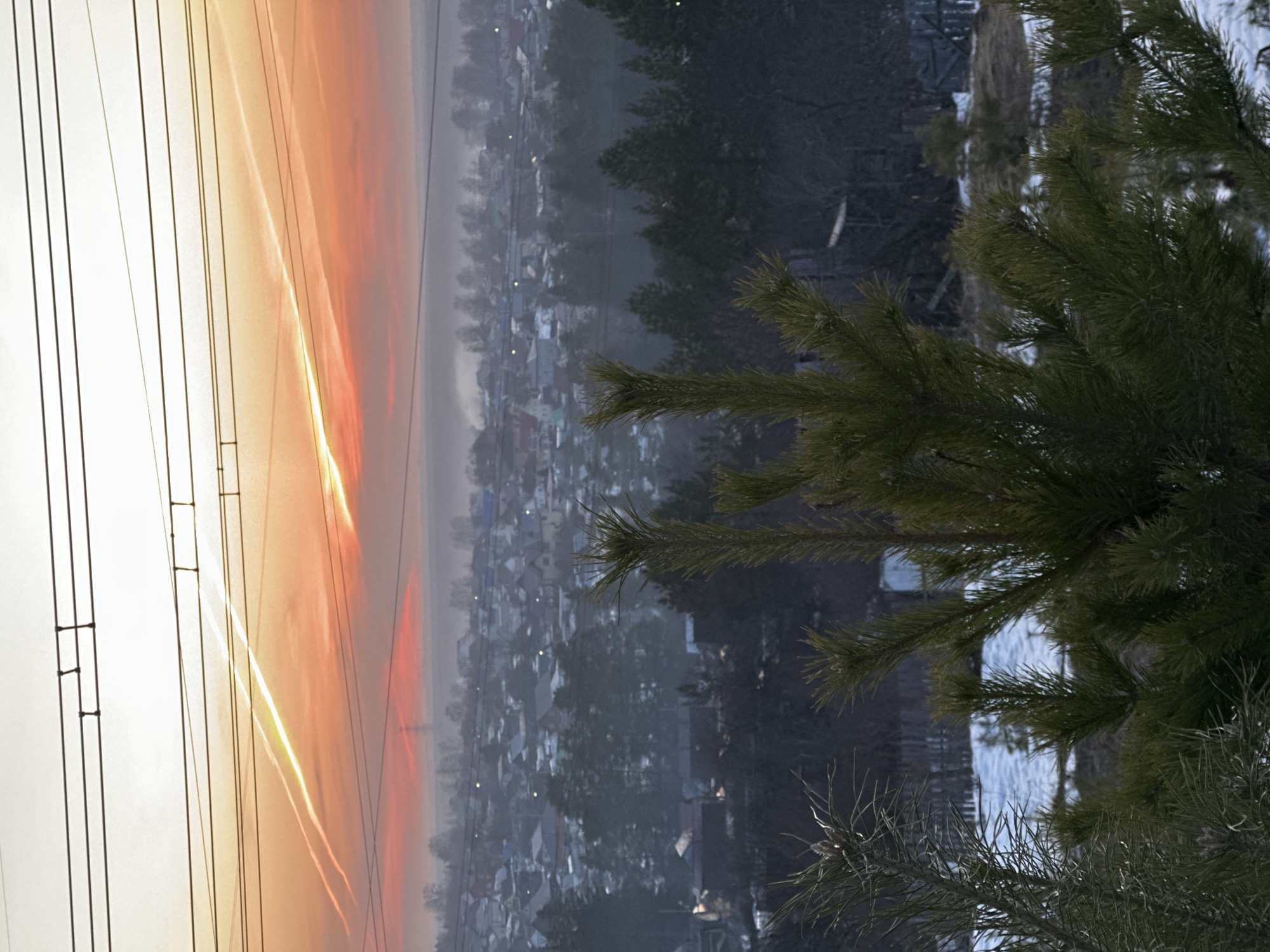 The image shows an elevated view of a residential area in Chaikovsky, Russia, at dawn or dusk. A stand of evergreen trees occupies the lower right corner of the frame. Across the middle ground is a dense neighborhood of small buildings. In the background, power lines intersect a sky tinted with warm hues, indicative of sunrise or sunset. Vapor trails cross the sky near the power lines. Snow covers the ground.