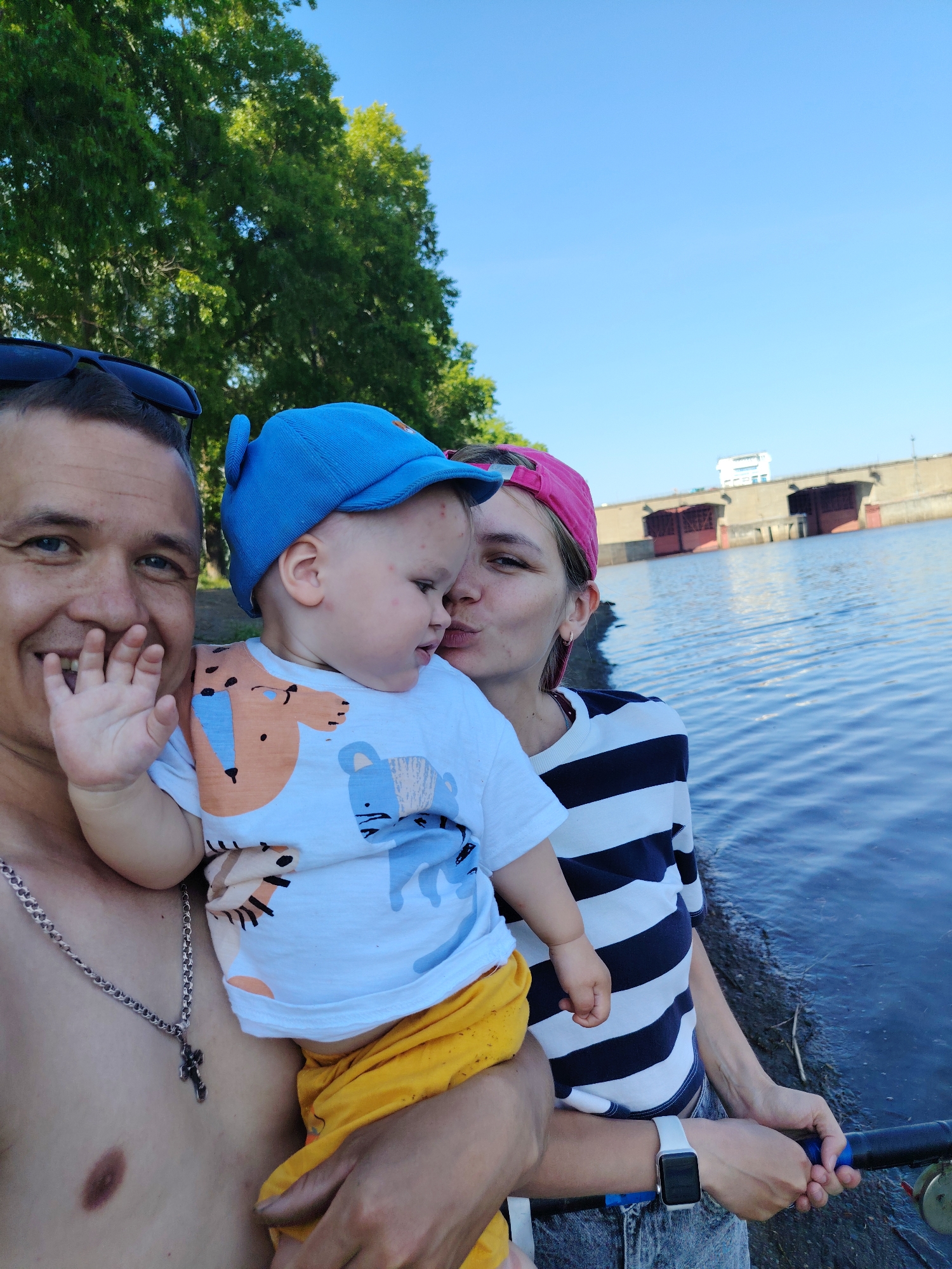 A family poses for a photo near a body of water in Chaikovsky, Russia. A man, bare-chested and wearing sunglasses atop his head and a silver cross necklace, smiles while holding a young child wearing a blue cap, a white shirt with animal designs, and yellow pants. A woman in a pink cap and a striped shirt kisses the child's cheek while holding a fishing rod. The background features trees, water, and a dam-like structure.