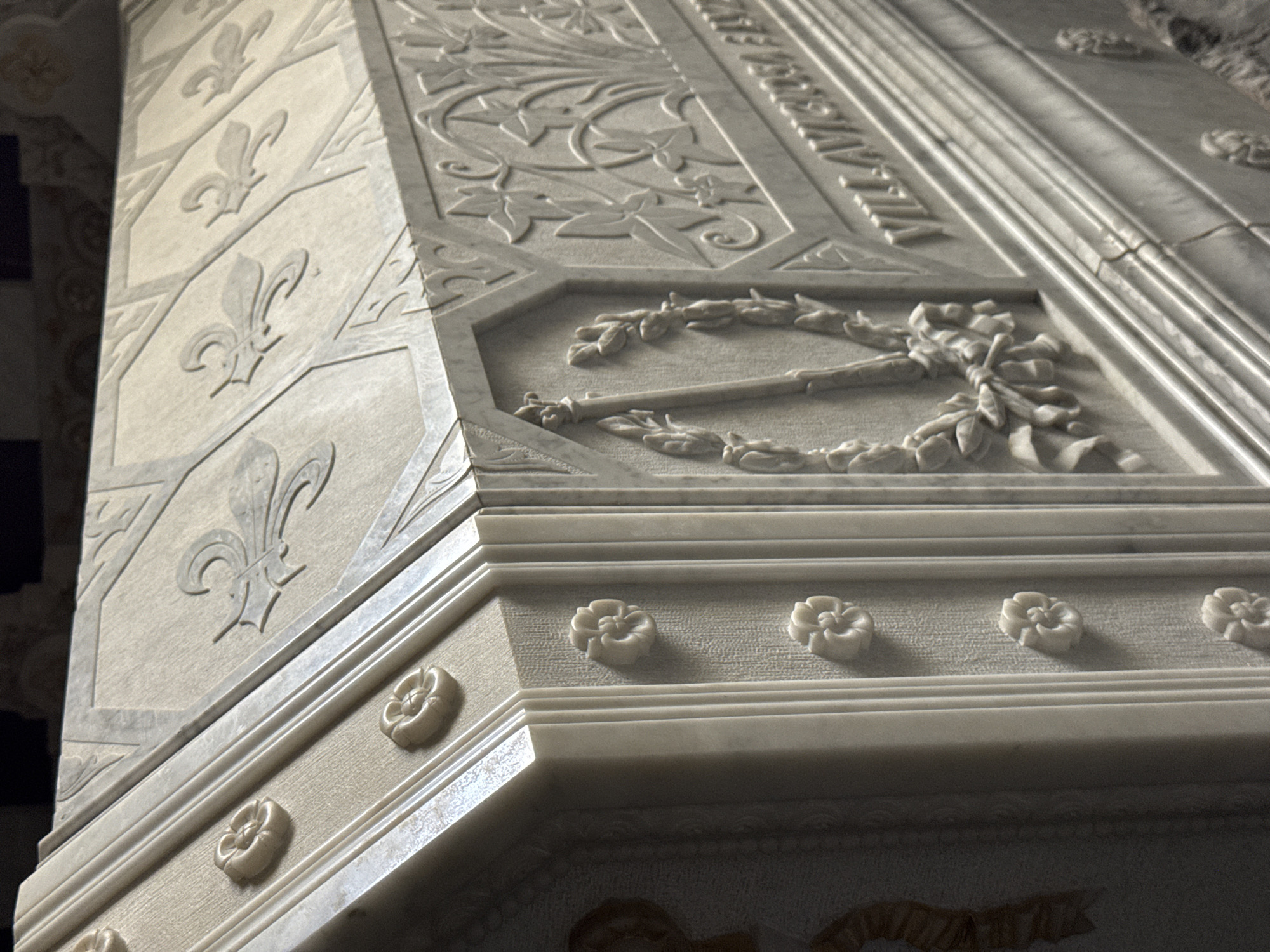 The image shows a close-up, angled view of a marble sarcophagus, likely a tomb, located in the Pantheon of Kings within the Monastery of El Escorial in San Lorenzo de El Escorial, Spain. The sarcophagus features intricate carvings, including floral motifs and what appear to be fleur-de-lis symbols on its lid. The side of the sarcophagus displays a relief carving of a wreath encircling a fasces, a symbol of authority. Partially visible text is etched into the marble on the side. The material is a light-colored marble, and the lighting highlights the detailed stonework. No people are visible in the image.