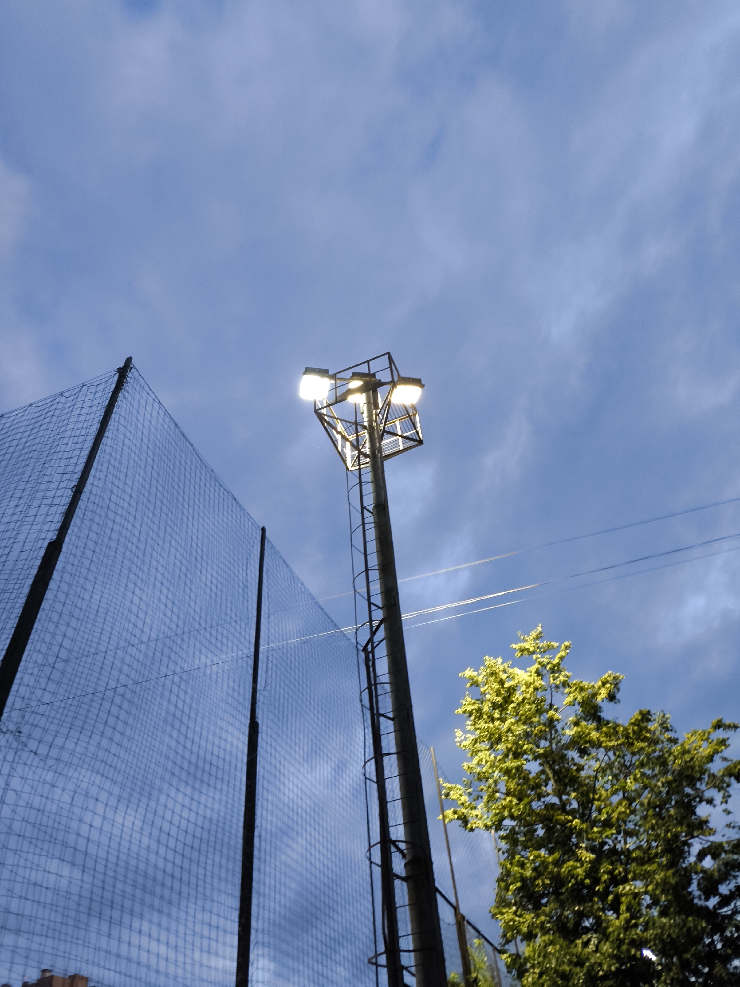 The media file shows a scene captured from a low angle, looking upwards towards a cloudy sky. The primary subjects are a tall, industrial-looking lamppost with bright lights illuminating the evening sky, a tall, dark fence made of netting, and a vibrant green tree with dense foliage. The lamppost is positioned to the right of the frame, featuring a metallic structure with multiple lights, suggesting it's likely for illuminating a sports field or outdoor area. A ladder is visible on the side of the lamppost. The fence, made of fine mesh netting, dominates the left and center of the image, with dark poles supporting it. The tree, situated on the right side of the frame, is bathed in light, highlighting its lush green leaves. The sky is a mix of blues and grays, indicating it is either dusk or dawn, with some wispy clouds. Faint contrails from an aircraft are visible in the sky. The overall impression is one of an outdoor recreational area at the end of the day, with artificial light beginning to take over from natural daylight. There is no visible text in the image.