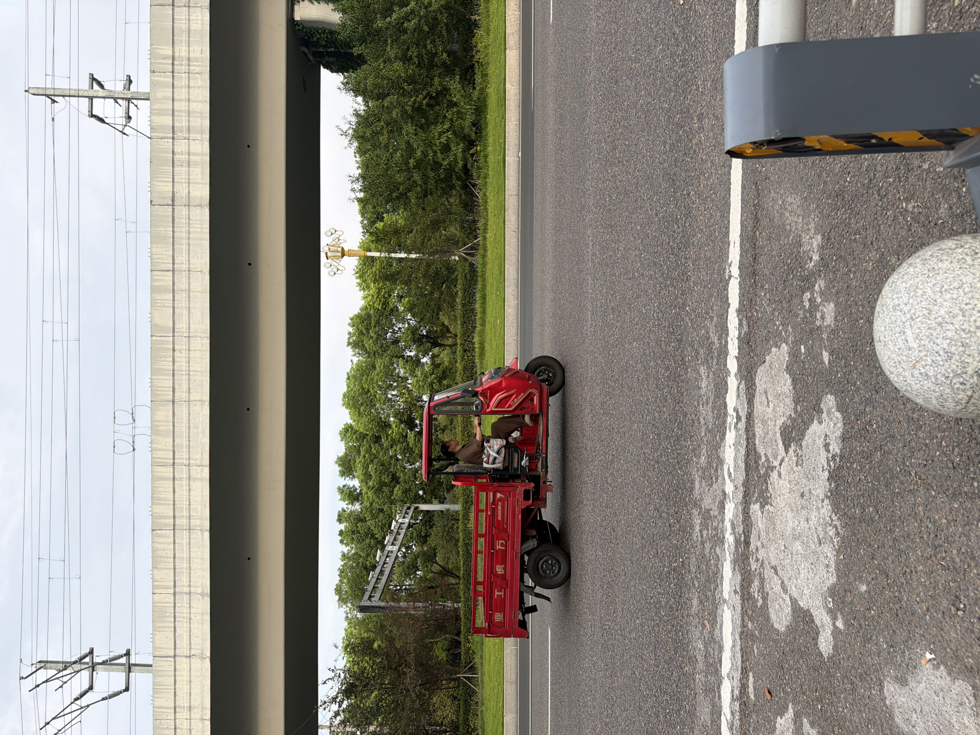 In Jiangjin, China, a person is driving a red, three-wheeled cargo vehicle on a paved road. The vehicle has a cab with a roof and a bed in the rear. The driver is wearing a brown shirt and pants and appears to be of Asian descent. The vehicle has some Chinese characters on the side. The road is bordered by a grassy median and a line of trees. In the background, a large overpass with overhead power lines is visible, suggesting an urban or suburban setting. The sky is overcast.
