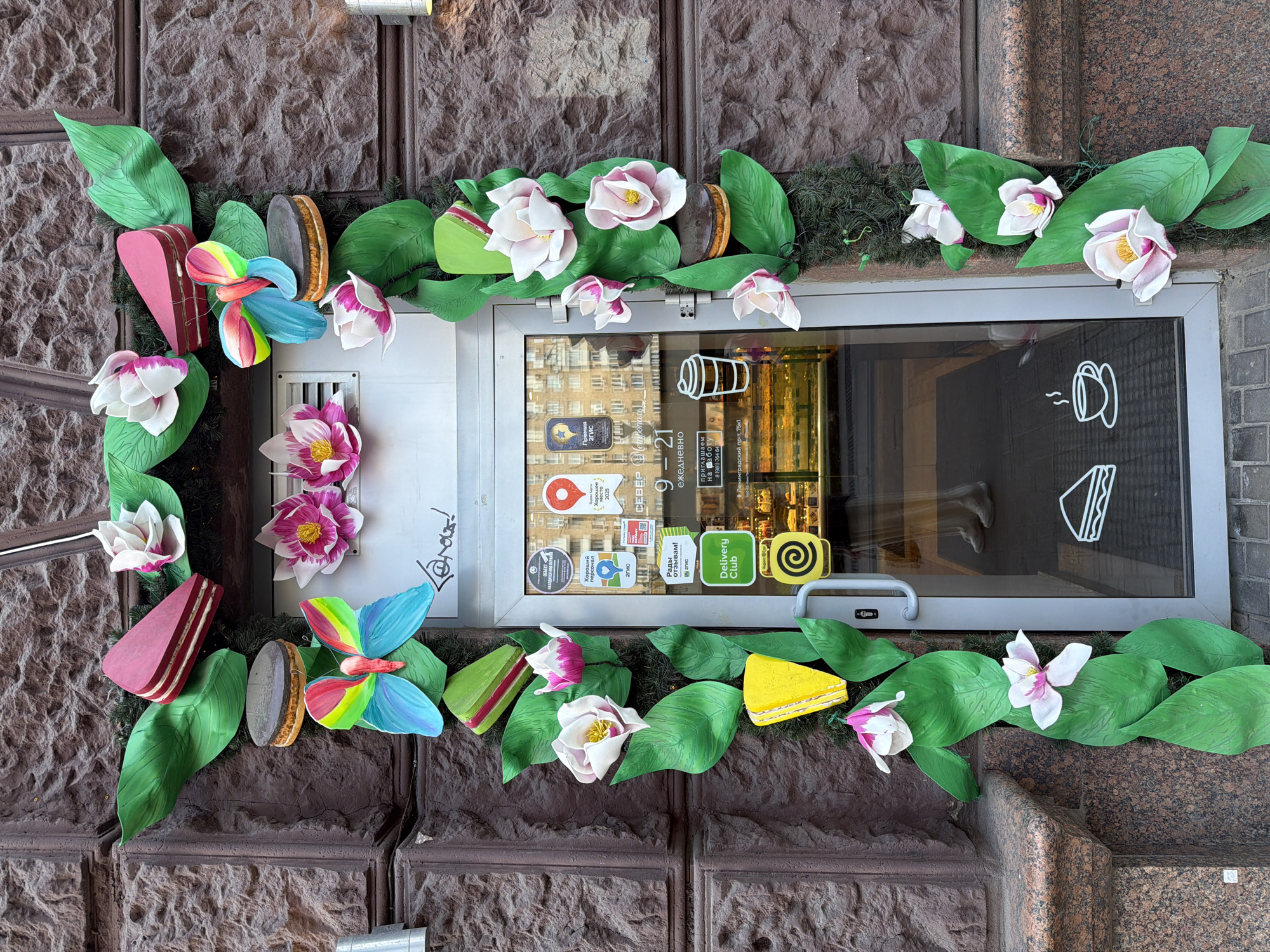 A storefront is decorated with artificial foliage and oversized food items. The facade of the building is made of brown brick. The storefront is a glass door with a metal frame, and it opens into a bakery visible through the glass. The decorations consist of large green leaves, pink and white flowers, brightly colored butterflies, and representations of cakes and cookies. The decorations are arranged in a garland-like fashion, framing the entrance. There are several stickers on the glass door, including those for "Delivery Club" and other services. A person's legs and feet, wearing light-colored shoes, are visible through the glass door, suggesting someone is inside the bakery. The overall scene depicts a whimsical and inviting storefront decoration, likely in a commercial area of Khimki, Russia.
