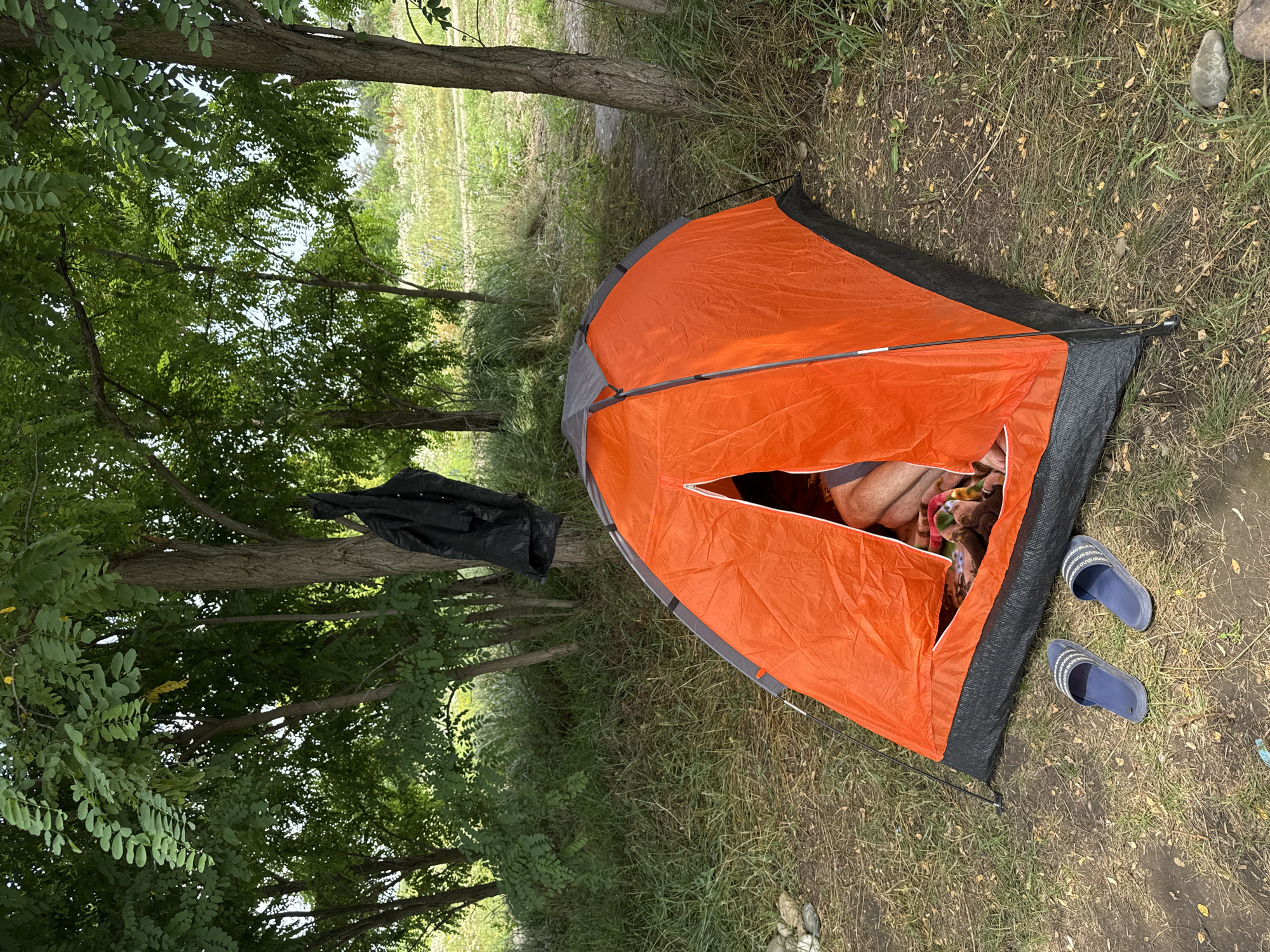 In Kochubeyevskoye, Russia, an orange tent is pitched under the shade of leafy trees. A person's legs are visible inside the tent, covered by a blanket. Two blue flip-flops rest on the ground outside the tent entrance. A black plastic bag hangs from a nearby tree branch. The ground is covered with grass, fallen leaves, and some stones. The background shows more trees and a field of dry grass and wildflowers.