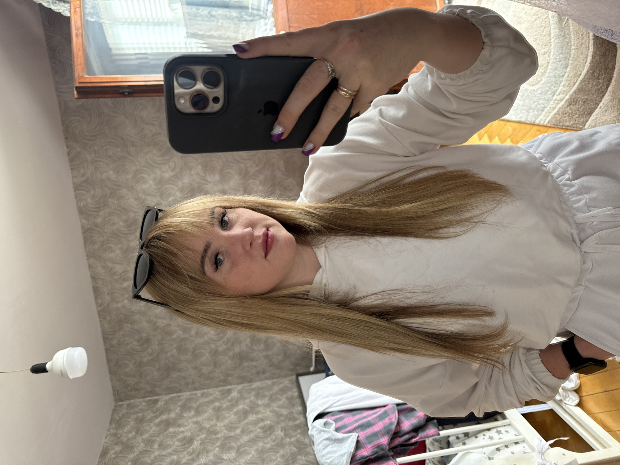 A woman with blonde hair and freckles is holding a black iPhone in front of her face, taking a selfie. She is wearing a white sweatshirt and has rings on her fingers, with purple and white nail polish. Sunglasses are perched on her head. The background includes a patterned wallpaper, a window, a light fixture, and what appears to be a partially visible crib with bedding. The location is presumably indoors, in what appears to be a residential setting. There is no information to confirm location in the city of Марковский, Russia.