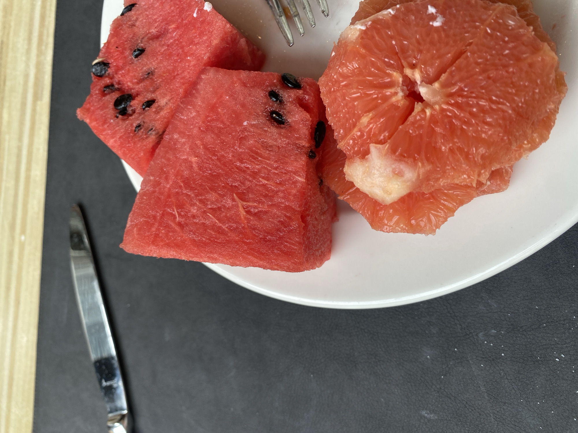 A close-up, overhead shot on a white plate shows slices of watermelon and grapefruit. The watermelon is cut into triangular wedges, revealing its red flesh and black seeds. The grapefruit is sectioned, showing its juicy, pink pulp. A fork is visible on the right side of the plate. A knife is also present in the upper left corner of the image, resting on a black surface. The background appears to be a light-colored wooden surface. The scene is set on a black textured table. The image does not depict any elements that would allow for the localization of Aksu, Turkey.