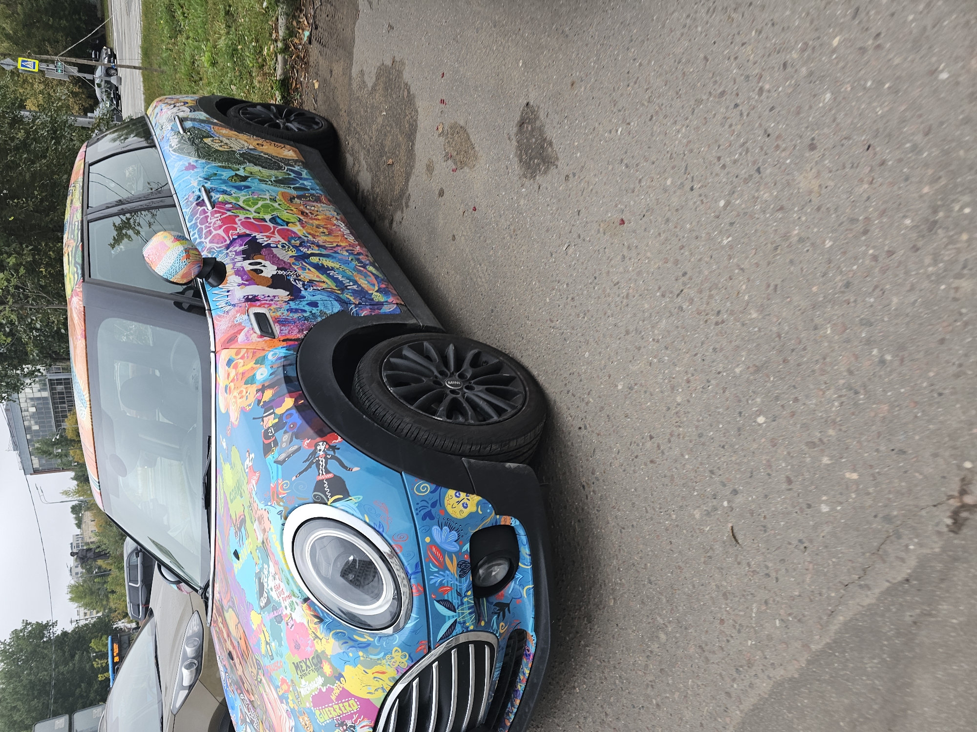 The image shows a Mini Cooper car with a vibrant, colorful, and complex graffiti-style wrap parked on an asphalt surface, possibly a street or parking lot, in Moscow, Russia. The car's body is covered in various cartoonish figures, skulls, abstract shapes, and text elements, creating a visually busy and artistic appearance. The car is viewed from its front three-quarter angle, with the driver's side facing the camera.
The environment appears to be urban. In the background, there are trees, buildings, and other vehicles. A light pole with a traffic sign is visible to the right of the car. The sky is overcast, suggesting a cloudy day. The asphalt ground shows some signs of wear and tear, including cracks and some dark wet patches, possibly from rain. There are also a few small reddish specks on the ground, which could be debris or fallen leaves.
No people are clearly visible in the image. The focus is on the uniquely decorated car.
