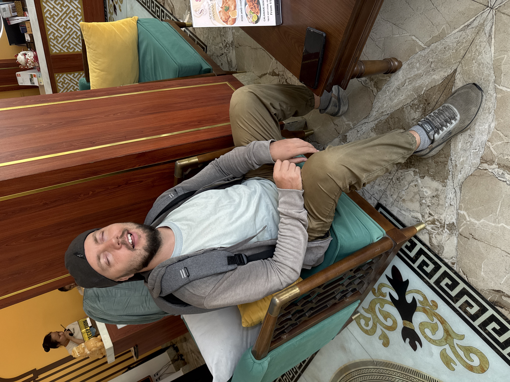 Here is a concise description of the image:
**Scene:** The image depicts a man reclining in a teal-colored chair with cushions in what appears to be a restaurant interior. He is wearing a grey hoodie, light blue t-shirt, and khaki pants. He is also wearing a black hat and grey sneakers.
**Objects:** A menu and a black phone sit on a wooden table next to the chair. The restaurant has a decorative floor with Greek-style patterns. There is a reception desk in the background, along with yellow and green cushions on a bench.
**Location:** Based on the menu visible on the table, the location is likely in Đà Nẵng, Vietnam.