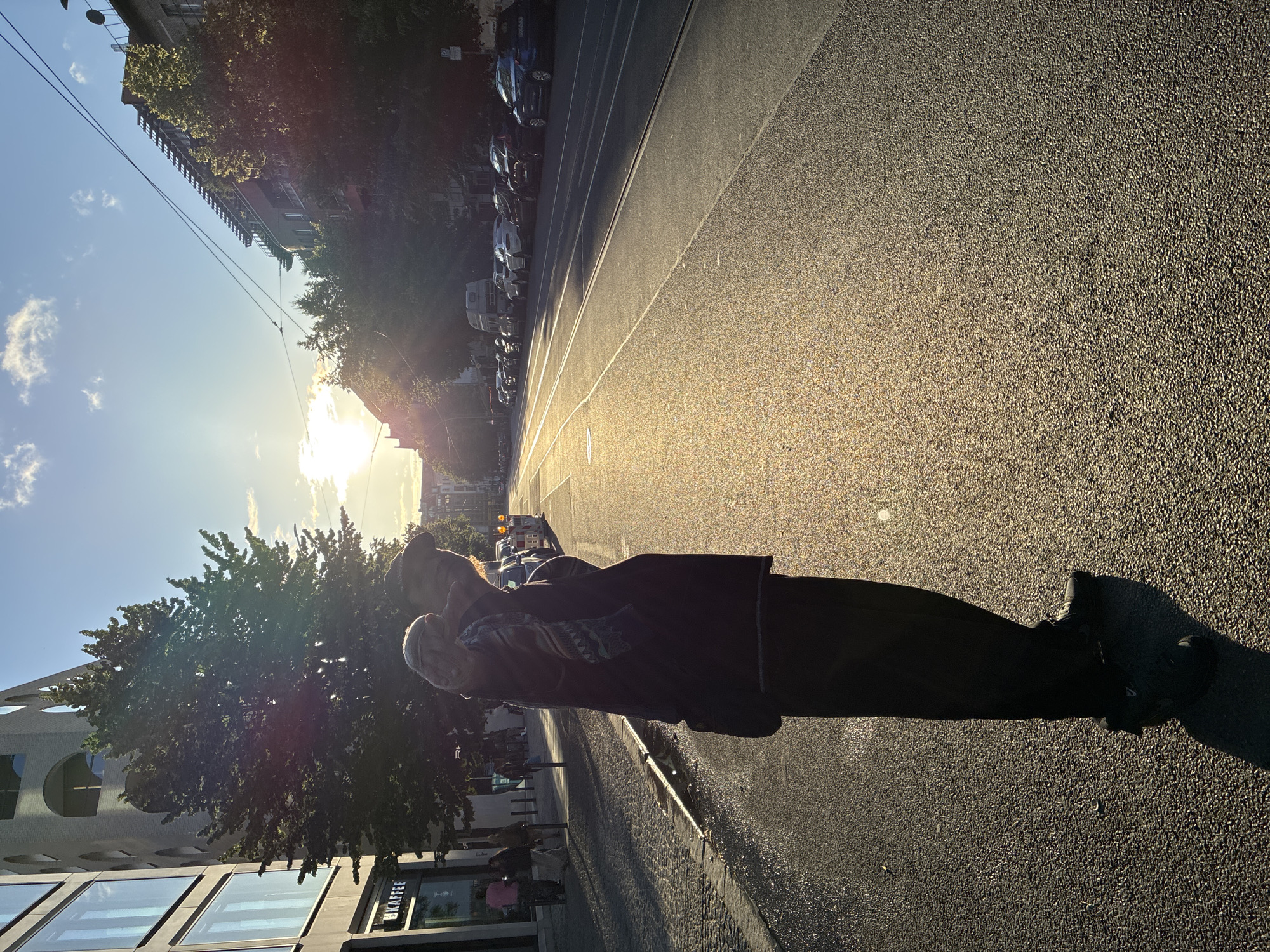 A full-body, low-angle shot captures a person standing on a street in Berlin, Germany, silhouetted against the golden light of a setting sun. The individual, wearing dark pants and a dark jacket with a patterned scarf draped over their shoulder, extends their right hand towards the camera, holding what appears to be a stuffed toy or blanket. The street, characterized by tram tracks, is wet and reflects the bright sunlight, creating a shimmering effect. Cars are parked along the right side of the street, and trees line the pavement on both sides. In the background, apartment buildings and a modern commercial building are visible, with overhead wires crisscrossing the sky. The scene suggests a warm, late afternoon or early evening setting in an urban environment.