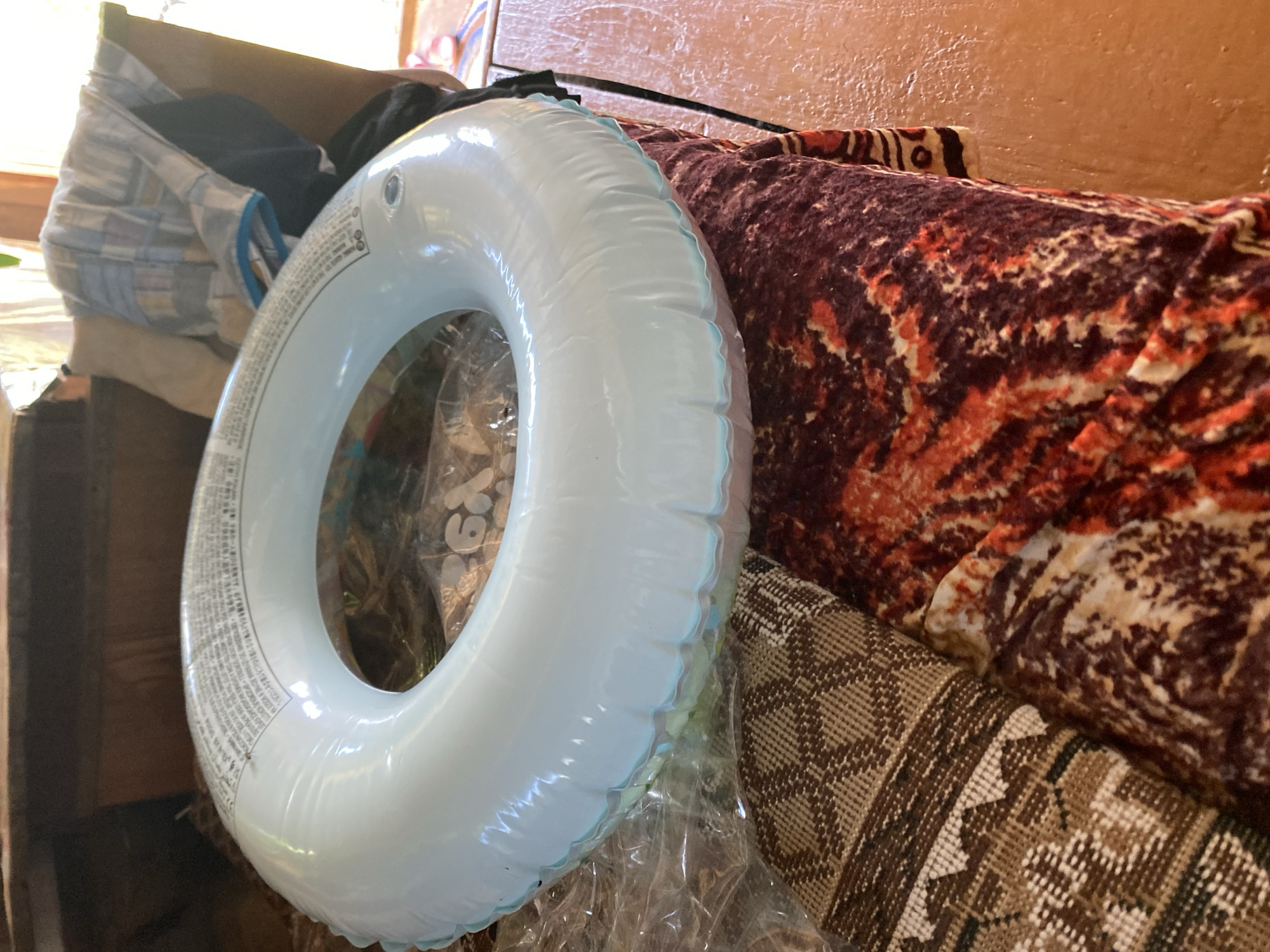 In the city of Maloarslangulovo, Russia, an inflatable blue swim ring rests on top of a pile of patterned blankets. The swim ring is positioned in the center of the image, partially obscuring the blankets beneath it. The blankets exhibit a mix of geometric and abstract patterns in brown, beige, and red tones. In the background, a wooden surface is visible, along with some folded clothing and what appears to be a window or doorway with natural light filtering in. The scene appears to be indoors, likely in a residential setting. No people are present in the image.