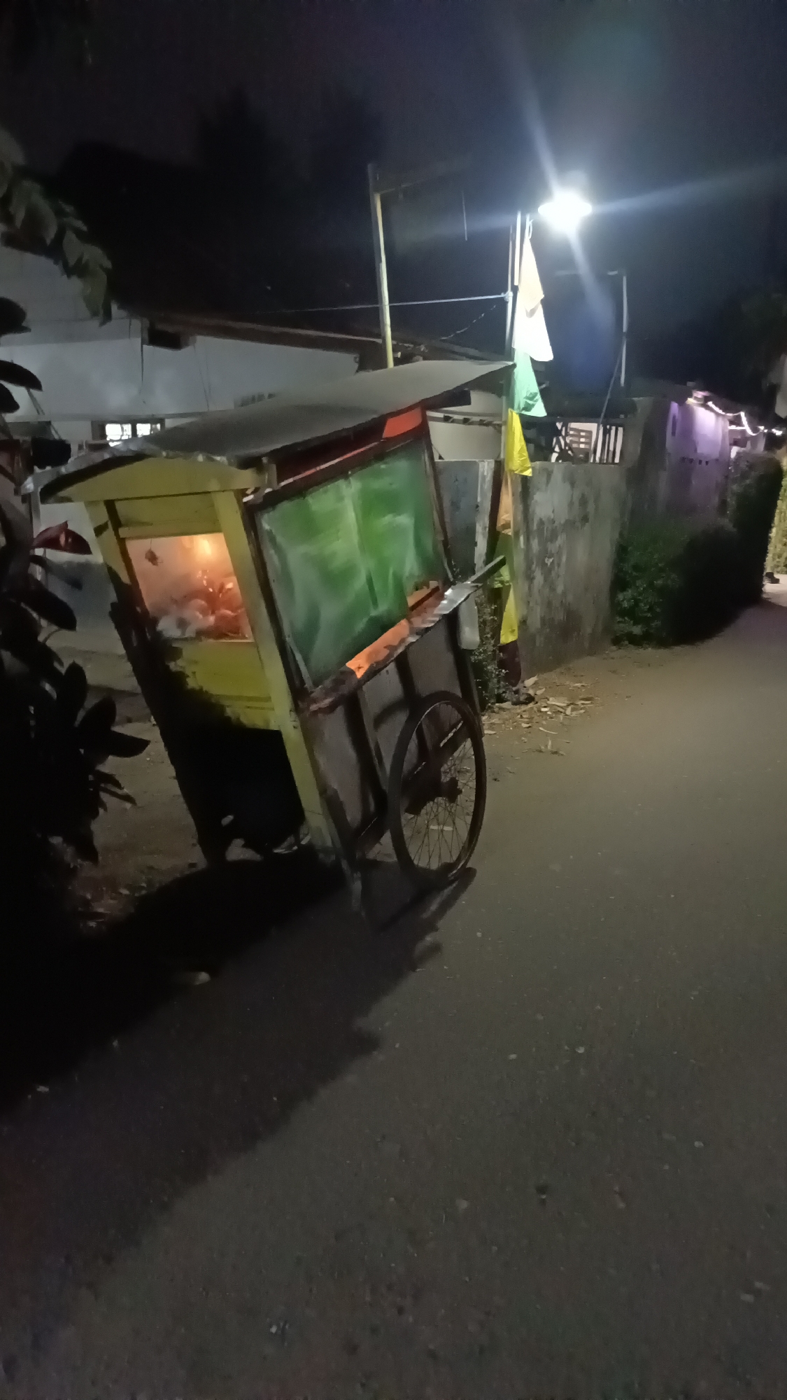 A food cart is parked on the side of a street in Kebumen, Indonesia at night. The cart is yellow with a green tarp covering part of its display area. The interior of the cart is lit, showing food items. The cart has one wheel visible and appears to be leaning. A tall pole with three colorful flags is erected behind the cart. Streetlights illuminate the scene. The background shows a wall and vegetation.