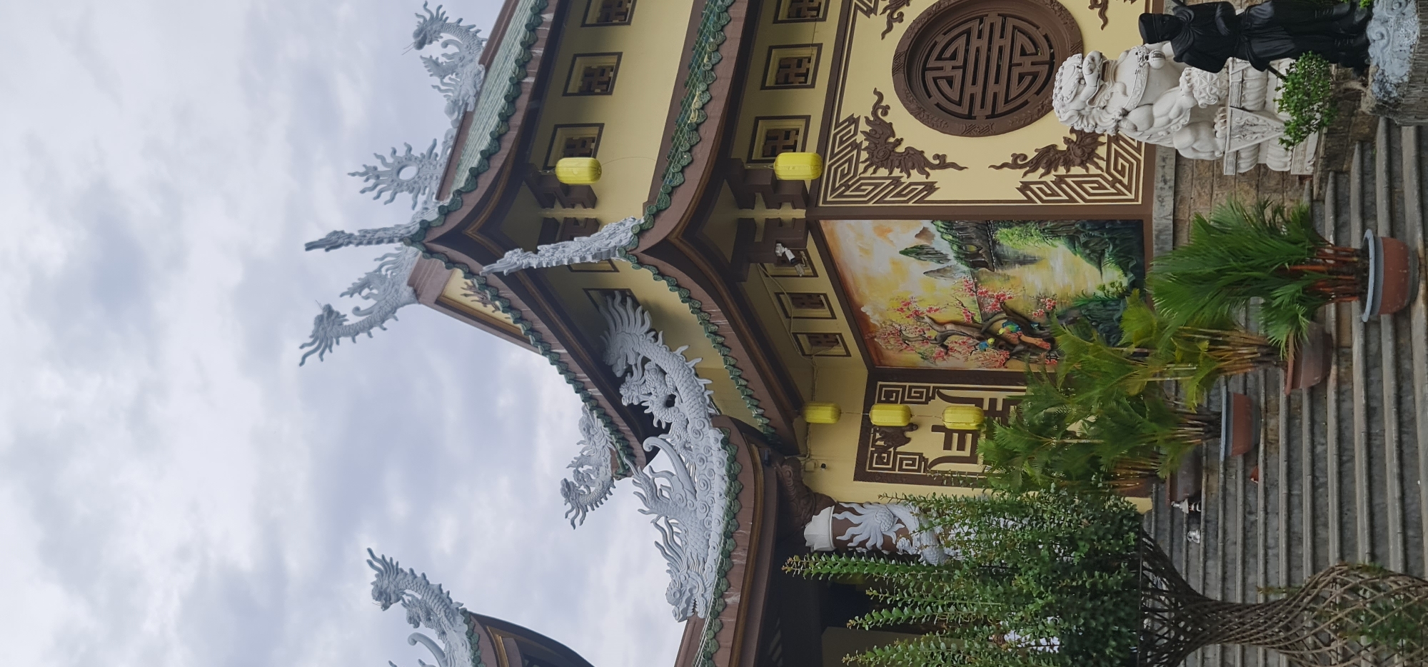 This image captures a close-up, upward-angled view of an elaborately decorated building, likely a temple or pagoda, in Da Nang, Vietnam, under an overcast sky. The architectural style is distinctly East Asian, characterized by multiple tiers of curving roofs with intricate green-tiled edges.
The most striking features are the numerous white dragon sculptures that adorn the eaves and roof corners. These dragons are detailed and appear to be leaping or climbing, adding a dynamic and traditional aesthetic to the structure. The building's walls are painted a warm yellow, accented with dark brown geometric patterns and frames. Several yellow lanterns hang from the eaves, providing small pops of color.
A large, vivid mural dominates a section of the wall, depicting a picturesque landscape with mountains, flowering trees—possibly cherry blossoms—and a serene body of water. Birds appear to be perched on the branches within the mural. Higher up on the wall, there's a circular wooden lattice window with an intricate design, and smaller square decorative panels.
On the right side of the image, a white stone statue, possibly a Foo dog or guardian lion, stands on a platform, partially obscured by lush green potted plants arranged on steps. A smaller, dark-colored human-like statue is also visible next to the white guardian. Another potted plant in a unique, woven conical planter is seen at the bottom right. The scene suggests a tranquil and culturally rich environment. The sky is a uniform grey, indicating cloudy weather during the daytime. No visible text is present.