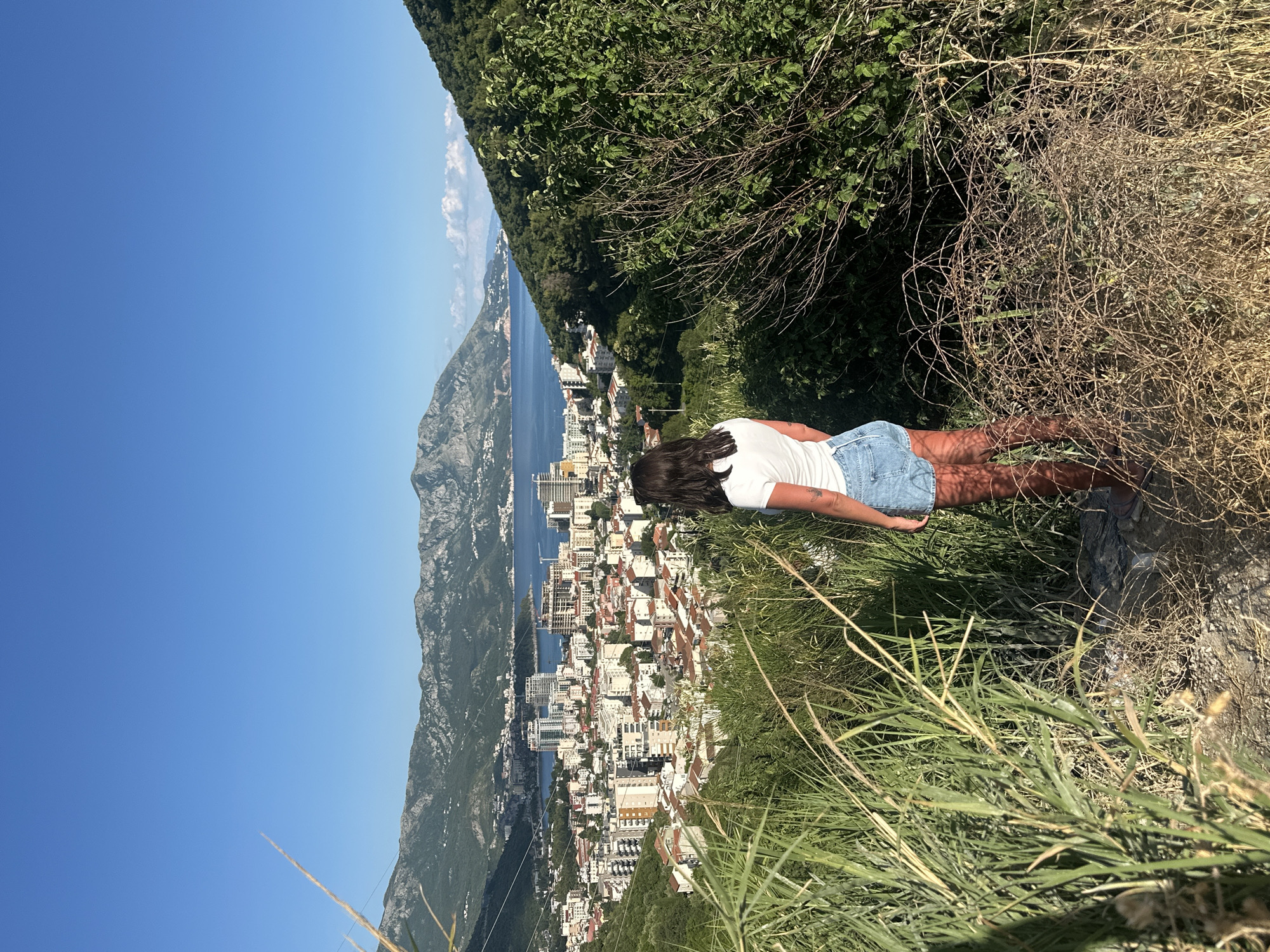 A young woman with dark hair stands facing away from the camera, looking out at a panoramic view of Budva, Montenegro. She wears a white t-shirt and denim shorts. The city sprawls below her, a densely packed urban landscape of buildings with red-tiled roofs and modern high-rises, nestled along the coast. In the distance, the clear blue Adriatic Sea meets a chain of majestic, tree-covered mountains under a bright, cloudless sky. The woman is positioned on a hillside covered in dense green vegetation and dry grasses, suggesting a natural overlook above the city.