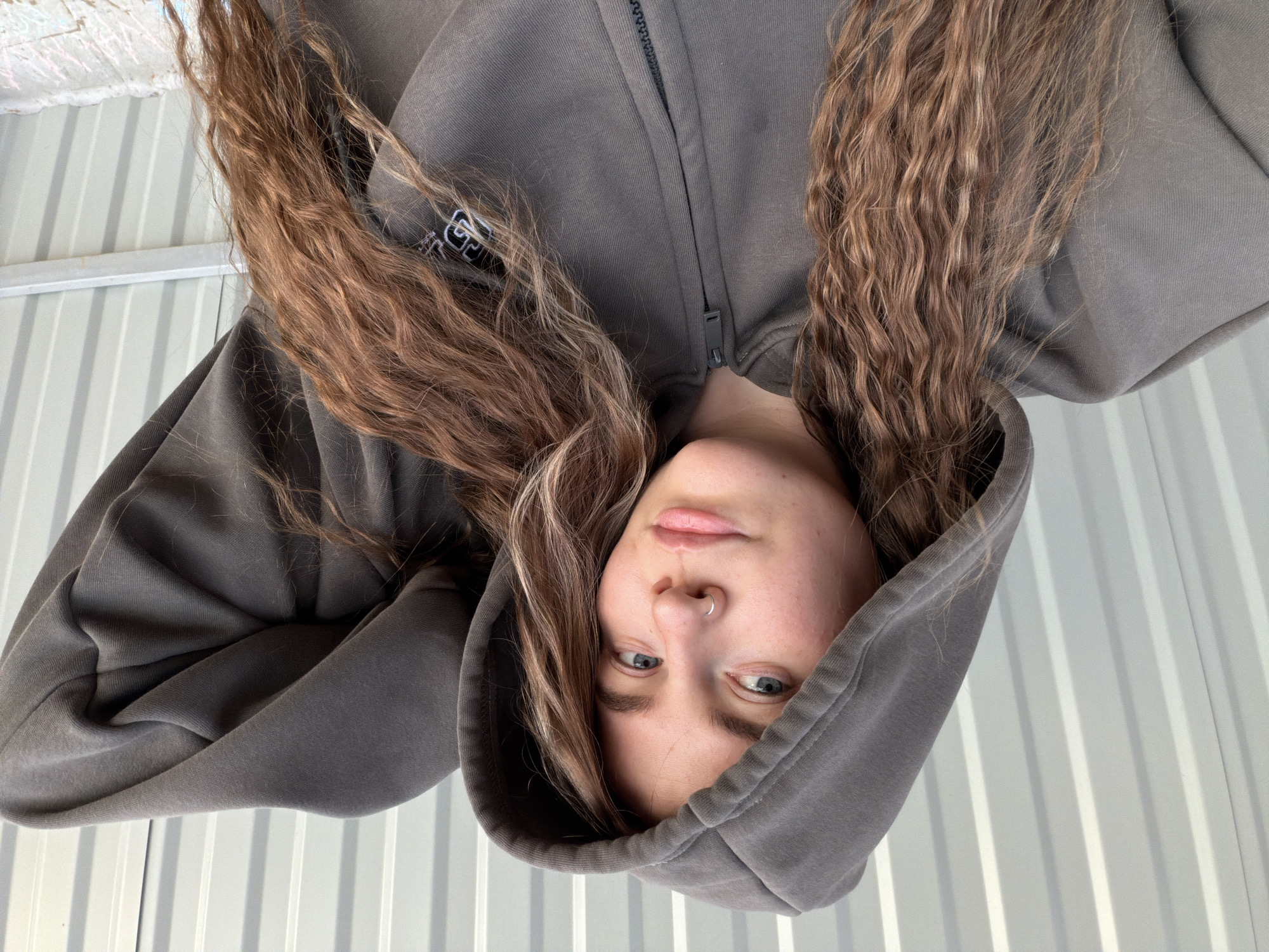 A young woman is photographed upside down, wearing a gray hooded sweatshirt that is zipped up. Her light brown curly hair hangs down, framing her face. She has blue eyes and a nose ring. The background is a white corrugated metal wall. The location, Chaikovsky, Russia, is based on information provided and is not discernable from the image itself.