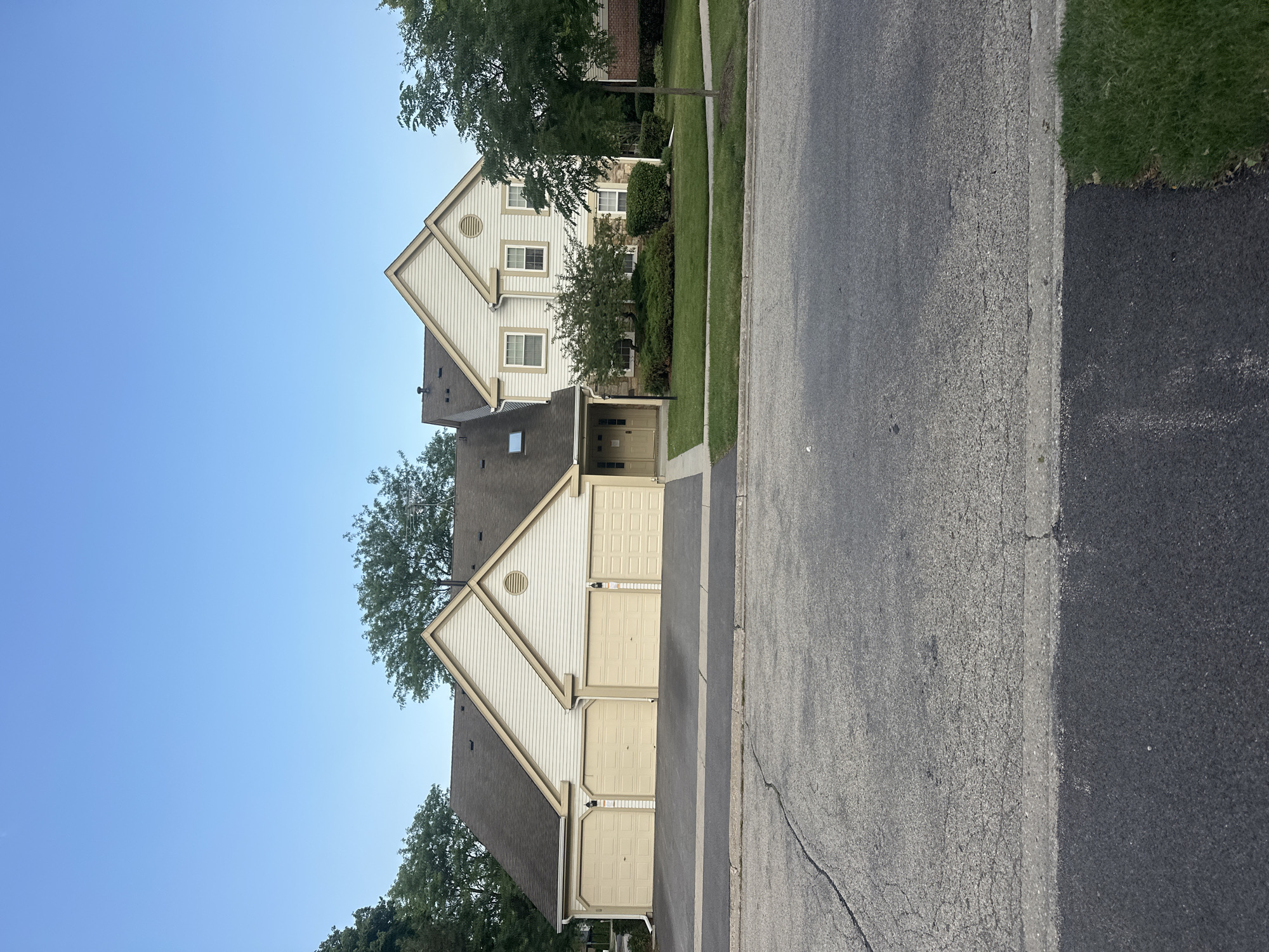 The image shows a tan, two-story townhouse complex with a dark brown roof and attached garages. The complex has four garage doors, each a light tan color. The townhouse on the right has white siding and windows, and a small tree and bushes in front. The sky is clear and blue. The scene is on a street in Schaumburg, Illinois, United States of America.