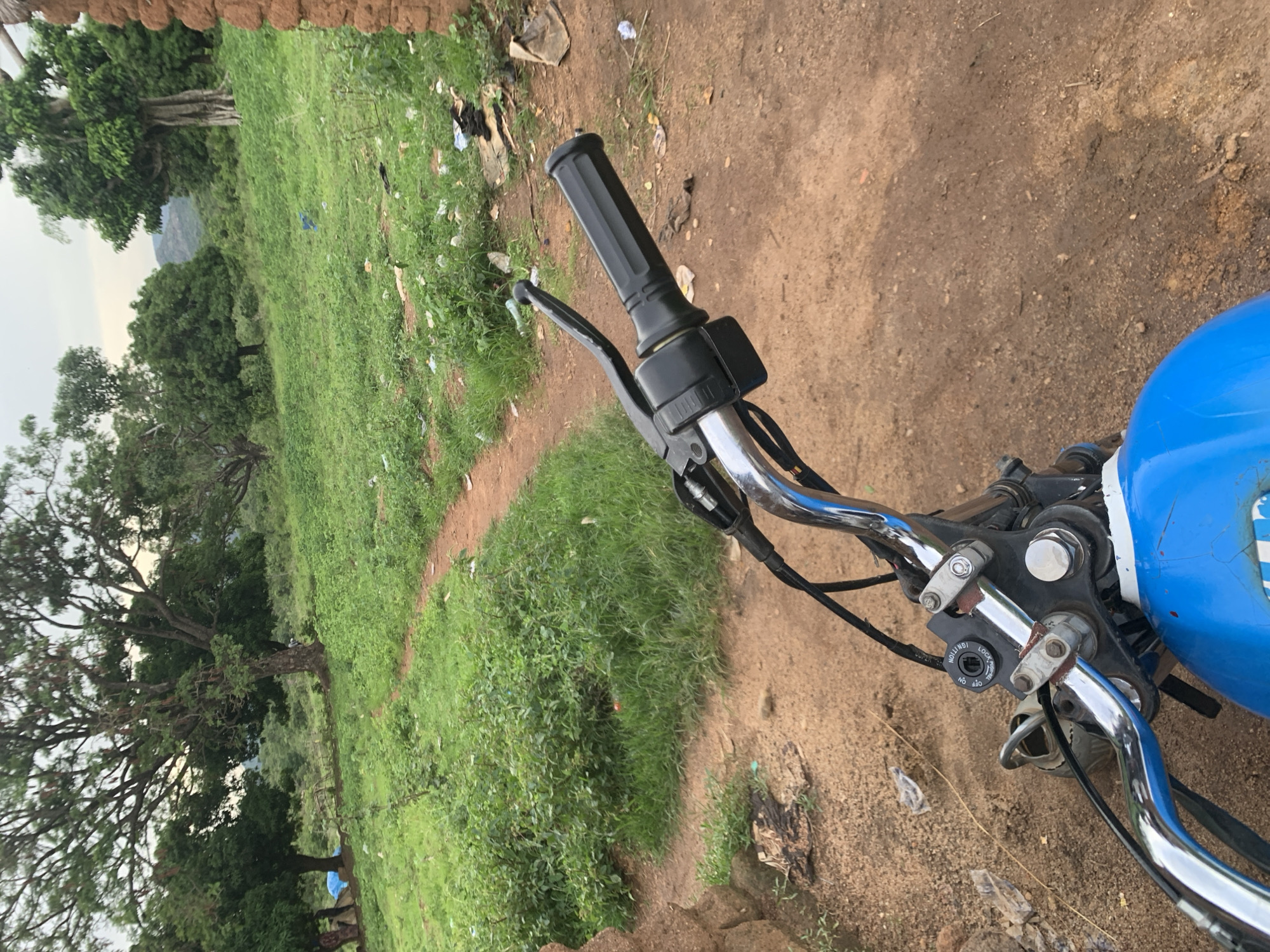 This media file is a first-person perspective shot from a motorcycle, looking forward and slightly to the left. The scene is outdoors in a rural setting, likely in Buga da Gindi, Nigeria, with a path winding through grassy terrain.
The foreground is dominated by the motorcycle's handlebars, which are chrome and black. A black grip is visible on the right side, along with a brake lever and a control unit. The blue fuel tank of the motorcycle is also visible on the right.
The background features a vibrant green, grassy landscape with scattered trees. Some of these trees are large and mature, with thick branches. In the distance, a hint of mountains or hills can be seen under an overcast sky. The ground beside the path is dry, dusty soil, with some scattered litter, including plastic bags and debris.
The time of day appears to be overcast, suggesting it might be morning or late afternoon, or possibly a cloudy day. The overall atmosphere is natural and somewhat unkempt, indicative of a rural or undeveloped area. No people are visible in the image.
The only visible text is on a circular control unit on the handlebar, which has settings for "IGNITION," "LOCK," "ON," and "OFF."