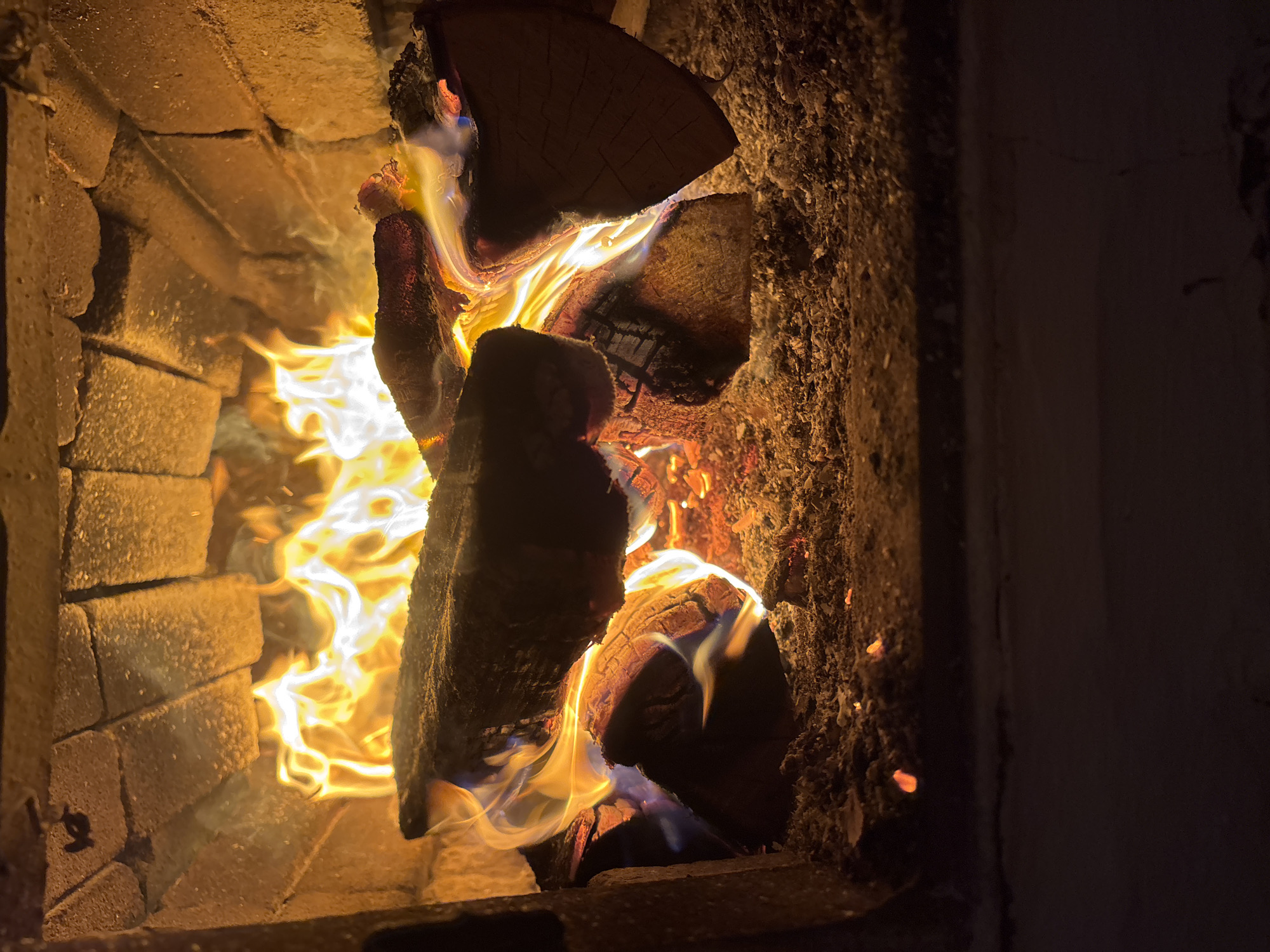 The image shows a close-up view of a burning fireplace filled with logs and flames. The fireplace is constructed of bricks, with the interior walls and floor covered in ash and glowing embers. The flames are a vibrant mix of yellow, orange, and white, creating a warm and inviting atmosphere. The logs are various sizes and shapes, with some appearing to be partially consumed by the fire. The background is dark, indicating the photo was taken in low light conditions. The scene does not provide any information about the city of Курган, Russia, or any specific location.