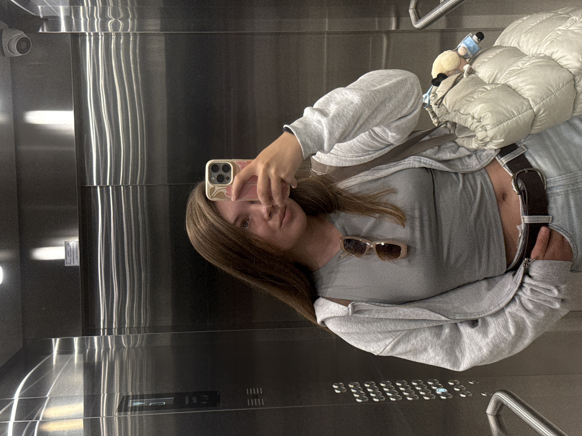 A young woman with long brown hair is taking a selfie in an elevator. She is wearing a grey crop top, a grey zip-up hoodie, and light blue jeans. A pair of sunglasses hangs from her hoodie, and she is carrying a beige puffy bag with a stuffed toy attached. The elevator walls are made of reflective stainless steel, and the floor buttons are visible on the left side. A security camera is mounted on the ceiling. The scene appears to be in a modern building, possibly a hotel or office building, within the city of Belgrade, Serbia.
