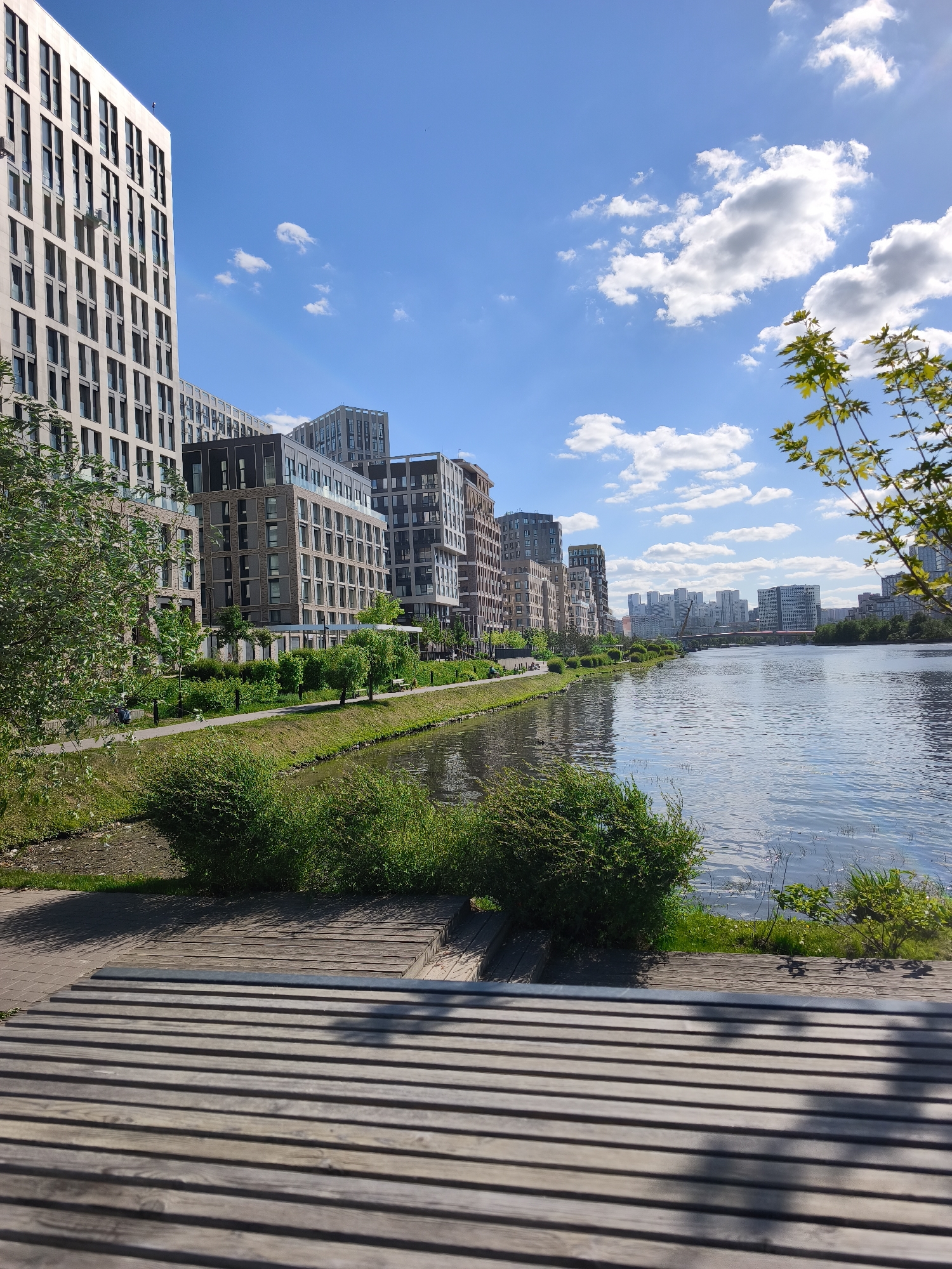 The image shows a cityscape along a river in Moscow, Russia. In the foreground is a wooden boardwalk-style seating area. The river reflects a clear blue sky with scattered cumulus clouds. Along the near bank of the river, there is a green strip with shrubs and trees. Behind this are modern residential buildings of varying heights and architectural styles. In the distance across the river, there are more buildings, including what appears to be a bridge. The weather is clear and sunny.