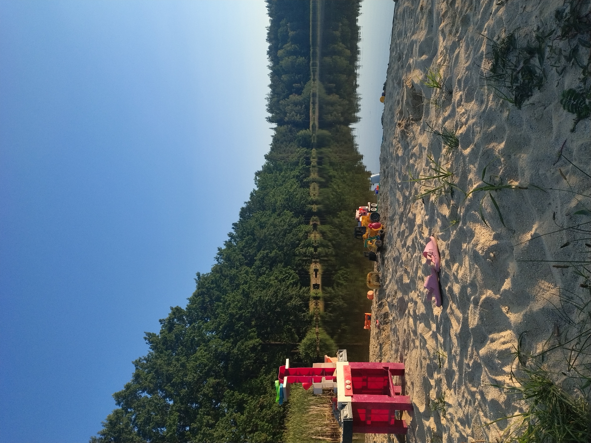 The image depicts a sandy beach area bordering a calm lake. The sky is a clear, bright blue. A dense line of green trees forms the background along the far shore of the lake, their reflections visible on the water's surface. In the foreground, on the sand, are various children's toys, including a pink play kitchen with plastic accessories, several toy construction vehicles (a yellow truck, a red and yellow truck, and a smaller yellow vehicle), and a pale pink pair of flip-flops. There are also some plastic balls and a white bucket scattered on the sand. The sand itself is light in color and shows indentations and small patches of green grass growing through it. The lighting suggests it is either early morning or late afternoon, with long shadows cast across the sand. The scene appears to be in a natural, outdoor setting, likely a recreational area.