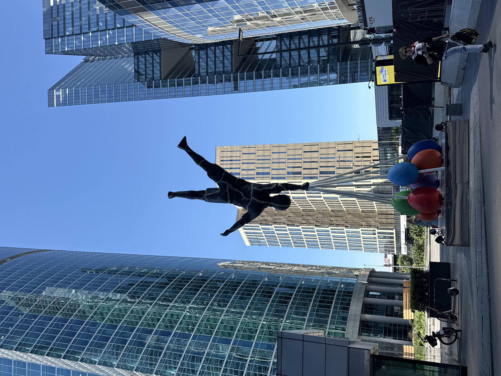 This image captures a modern urban scene in the city of Khimki, Russia, under a clear blue sky. Dominating the center is a dark, textured sculpture of a human figure performing a handstand, balanced on several angled, silvery stilts. Below the figure, at the base of the stilts, are several large, brightly colored, egg-shaped objects in red, green, blue, purple, and orange.
To the left, a tall, curvilinear skyscraper with a reflective greenish-blue glass facade rises. The facade exhibits intricate reflections of other buildings and the sky. Its lower section features large, cylindrical support columns. To the right of the central sculpture, another complex of modern glass skyscrapers, angular in design and reflecting the blue sky, extends upwards. A third building, lighter in color with repetitive vertical window patterns, stands directly behind the sculpture in the mid-ground.
In the right foreground, a person with light, curly hair, wearing sunglasses, a dark t-shirt, light jeans, and dark sneakers, walks to the right, carrying a dark bag and possibly a phone. A white and red garment is draped over their shoulders.
At ground level, in the left foreground, a bicycle is parked at a bike rack next to an electric scooter. The ground is paved with light-colored flagstones. In the mid-ground, behind the sculpture and the person, a yellow sign displaying Cyrillic text "Сбербанк Путешествия" (Sberbank Travel) is visible, along with black construction barriers and a white truck featuring a "GTS" logo. Low green hedges and other sparse vegetation are also present in the background. The scene is brightly lit by natural sunlight.