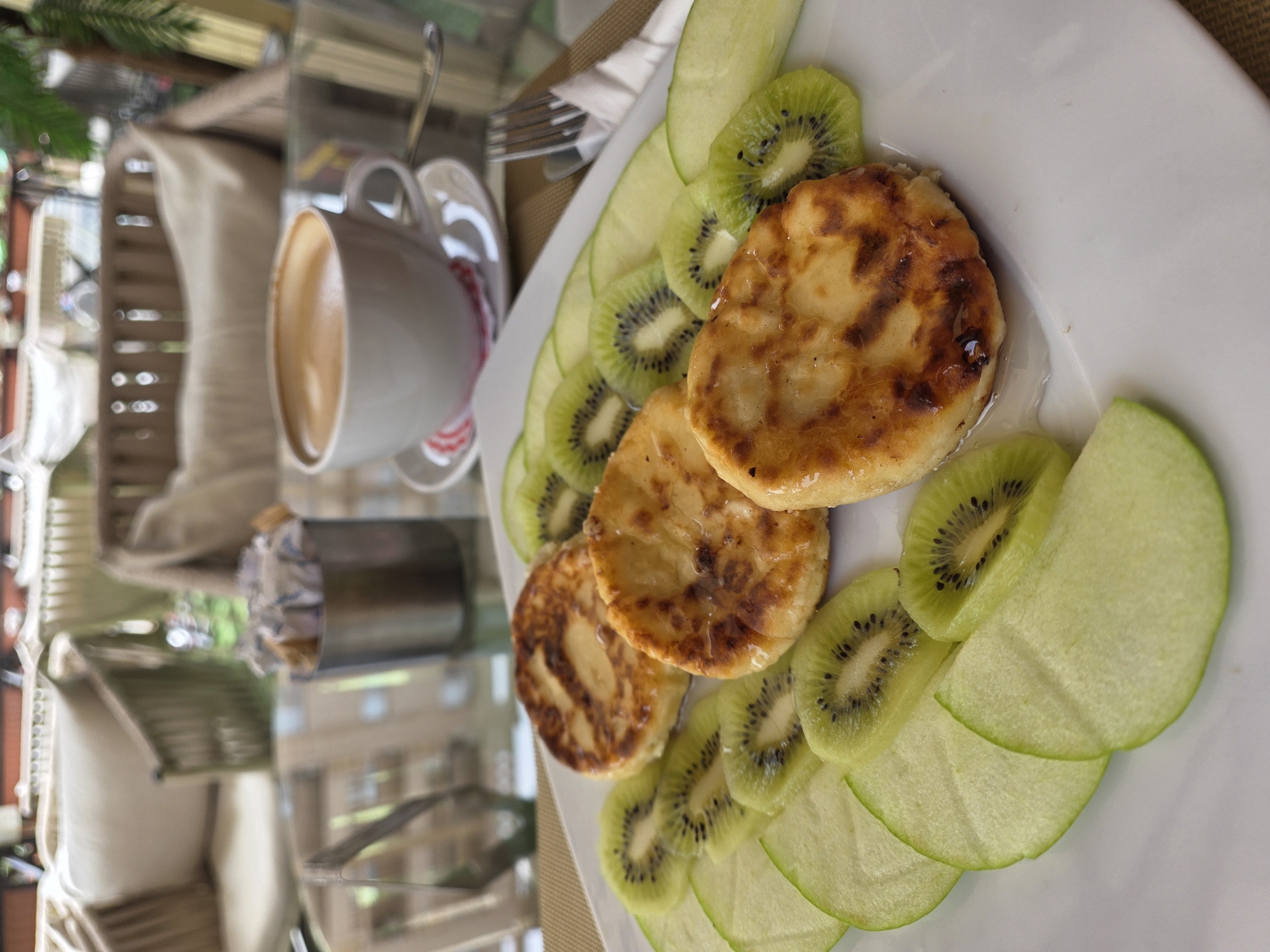 The image depicts a table at an outdoor cafe in Hurghada, Egypt. A white plate is centered on the right side of the frame, containing three golden-brown syrniki (small fried quark pancakes), garnished with slices of kiwi and green apple. To the left of the plate is a white ceramic cup filled with coffee, set on a saucer with a spoon and fork alongside. The table is made of glass and reflects the surroundings, including a glimpse of a building outside. Seating with cushions is partially visible in the background. The table is covered with a brown placemat.