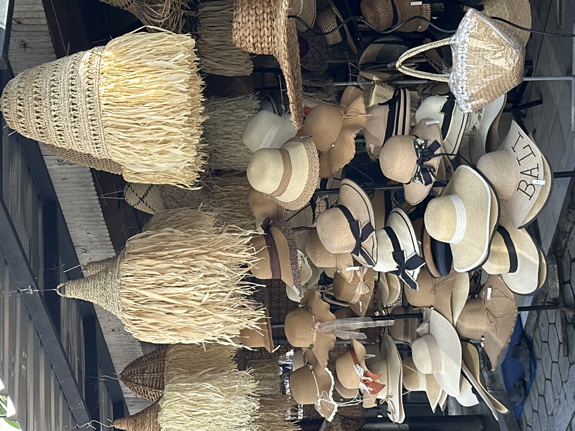 This image depicts a street stall in North Kuta, Indonesia, showcasing a variety of woven hats and lampshades. The stall is covered by a slatted roof, and an assortment of straw hats are displayed on tiered racks. Many of the hats have wide brims and are decorated with ribbons, bows, or lace. Above the hats, several large, conical lampshades made of woven straw with fringe detailing are hung. To the right, a woven straw handbag with a decorative trim and plastic covering is on display. The ground is paved with hexagonal tiles, and a pair of dark grey slippers are visible on a blue mat in the lower left corner. The scene is brightly lit, suggesting daytime. There are no people visible in the frame.