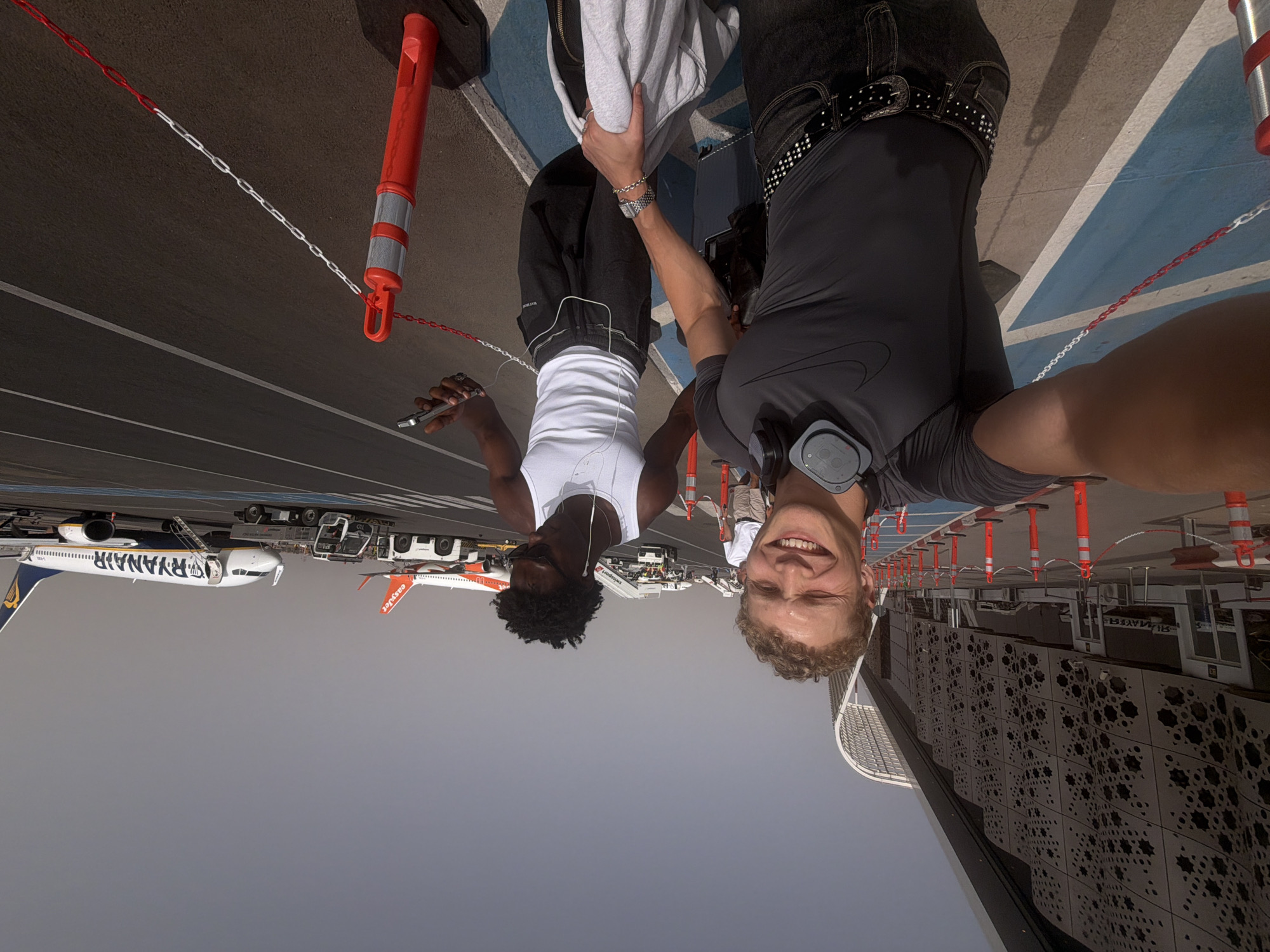 The image depicts a sunny day at Marrakesh Menara Airport in Marrakesh, Morocco. Two young men are posing for a selfie on the tarmac. The man in the foreground, with fair skin and curly blonde hair, wears a dark grey Nike t-shirt, dark jeans with a studded belt, and headphones around his neck. He is smiling and holding a camera. The second man, with dark skin and black curly hair, is wearing a white tank top and dark sweatpants. He is looking down at his phone, which he holds in his right hand, and has earbuds in. In the background, a Ryanair airplane is visible on the tarmac, as is part of an easyJet plane. The modern airport terminal building with its distinctive geometric facade is to the left. Red and white safety barriers and cones line the edge of the tarmac. The sky is clear and the overall environment appears to be an active airport setting.