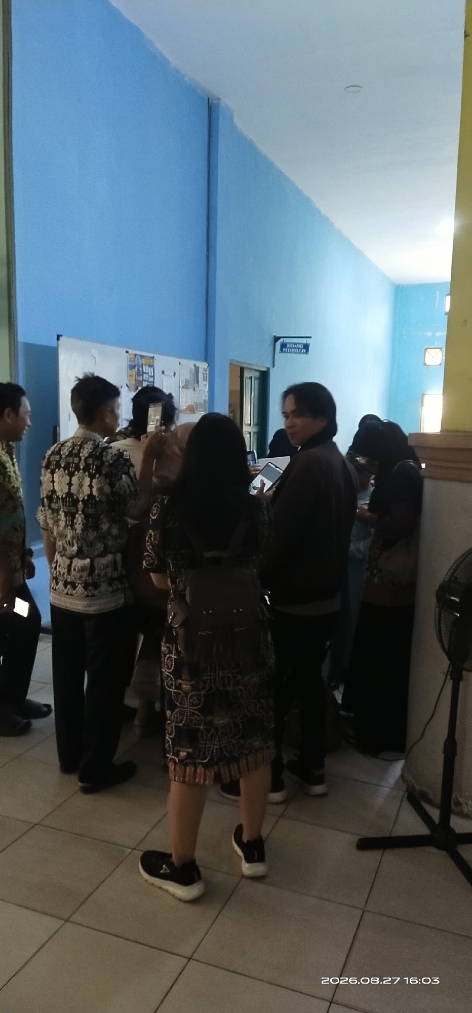 In Tamiang Layang, Indonesia, a group of individuals is gathered in what appears to be a hallway or corridor with light blue walls. The flooring is tiled in a light beige color. On the wall to the left, there is a bulletin board covered with papers. A doorway is visible on the right, with a sign above it that reads "BIDANG PETERNAKAN," indicating the Animal Husbandry Department.
Several people are present. On the far left, a person in a dark patterned shirt and black pants is visible from the shoulders down, holding a white object that may be a phone. Next to them, a person wearing a black and white patterned batik shirt and black pants stands facing away from the camera. In the center, a person with long dark hair, wearing a dark jacket and dark pants, is holding a tablet and looking to their right. To their right, a person with long dark hair, wearing a patterned dress and a backpack, is facing away. Further to the right, several other individuals are partially visible. A black standing fan is positioned on the far right of the frame, with its base and some of the blades visible. The timestamp "2026.08.27 16:03" is present in the bottom right corner of the image.