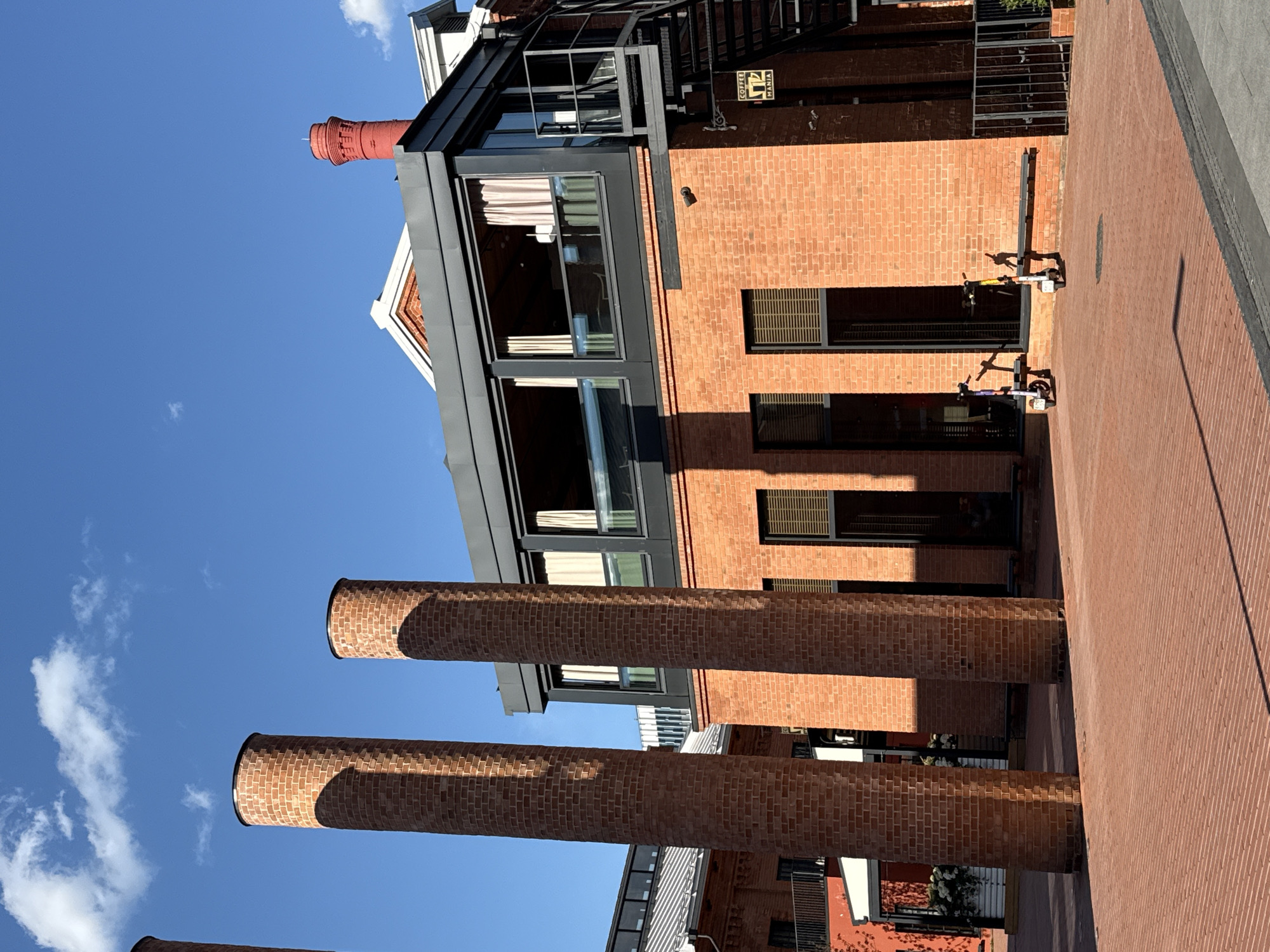 The image displays an urban scene in Moscow, Russia, dominated by red brick architecture under a bright blue sky. The perspective is angled, looking up at the buildings and tall structures.
The main subjects are several tall, cylindrical brick chimneys in the foreground and midground, casting long shadows. Behind them, a multi-story building constructed of red bricks features large, dark-framed windows, some with curtains visible inside. This building also has a modern-looking black metal balcony and external staircase structure. A smaller, decorative red brick chimney is visible on its roof. Another brick building to the right has smaller windows with grilles.
The ground is paved with uniform red bricks, matching the building materials and creating a cohesive aesthetic. Two electric scooters are parked on this brick pavement near the building, indicating a contemporary urban setting.
No explicit human activity is taking place, but the parked scooters suggest potential transportation or leisure in the area.
Notable details include the bright, clear blue sky with a few scattered white clouds, indicating fair weather. The strong, angular shadows cast by the chimneys and buildings suggest it is daytime, likely mid-morning or afternoon, with the sun at a significant angle.
A yellow, square-shaped sign with the text "COFFEE MANIA" is affixed to the brick wall of one of the buildings, featuring an illustration of a steaming coffee cup. The overall scene suggests a repurposed industrial or historical architectural space, now serving modern uses.
