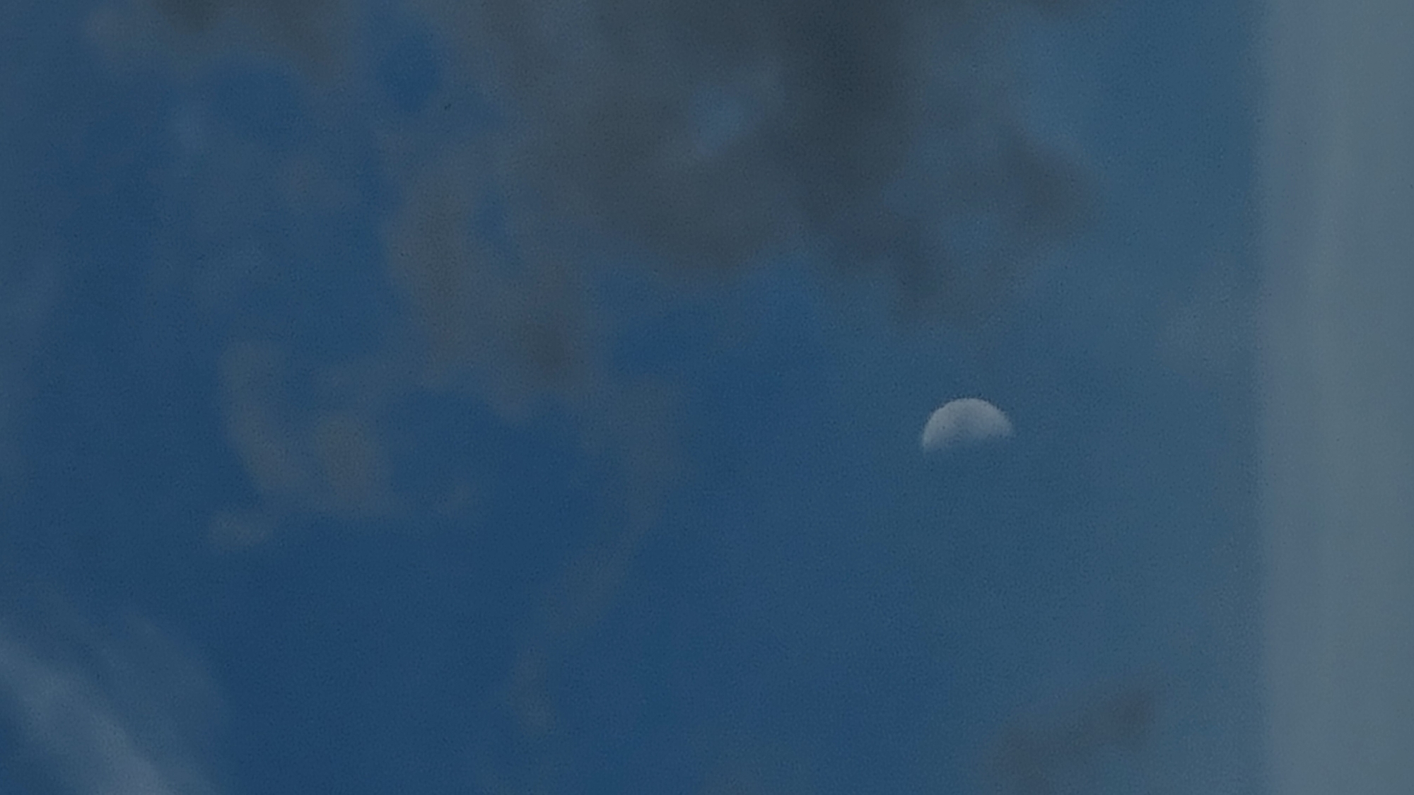A partially illuminated moon is visible in a dark blue sky. The moon appears as a crescent shape, with its illuminated portion facing towards the left. There are faint clouds scattered across the sky, appearing as lighter grey smudges against the darker blue background. The image is a vertical shot, and the bottom portion of the frame shows a lighter, indistinct area that could be clouds or a distant landscape. There are no people or man-made structures clearly identifiable in the image. The scene is consistent with an outdoor view in Vysokoe, Russia, during twilight or nighttime conditions.