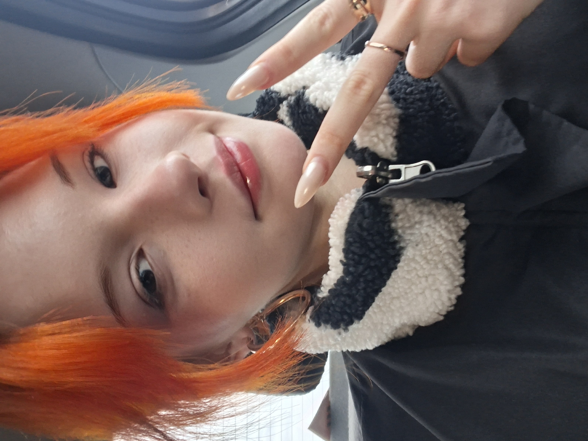 A selfie of a person with orange hair, a fair complexion, and light pink lips, is taken at an oblique angle. The person is wearing a black jacket with a white, sheepskin-like collar. The person has long, almond-shaped fingernails, and wears a gold ring on the index finger. The person is making a 'V' sign with their fingers. A gold hoop earring is visible in the left ear. The background is blurry, but the window of a vehicle can be discerned. It is impossible to determine the location in the city of Chaikovsky, Russia from this image.