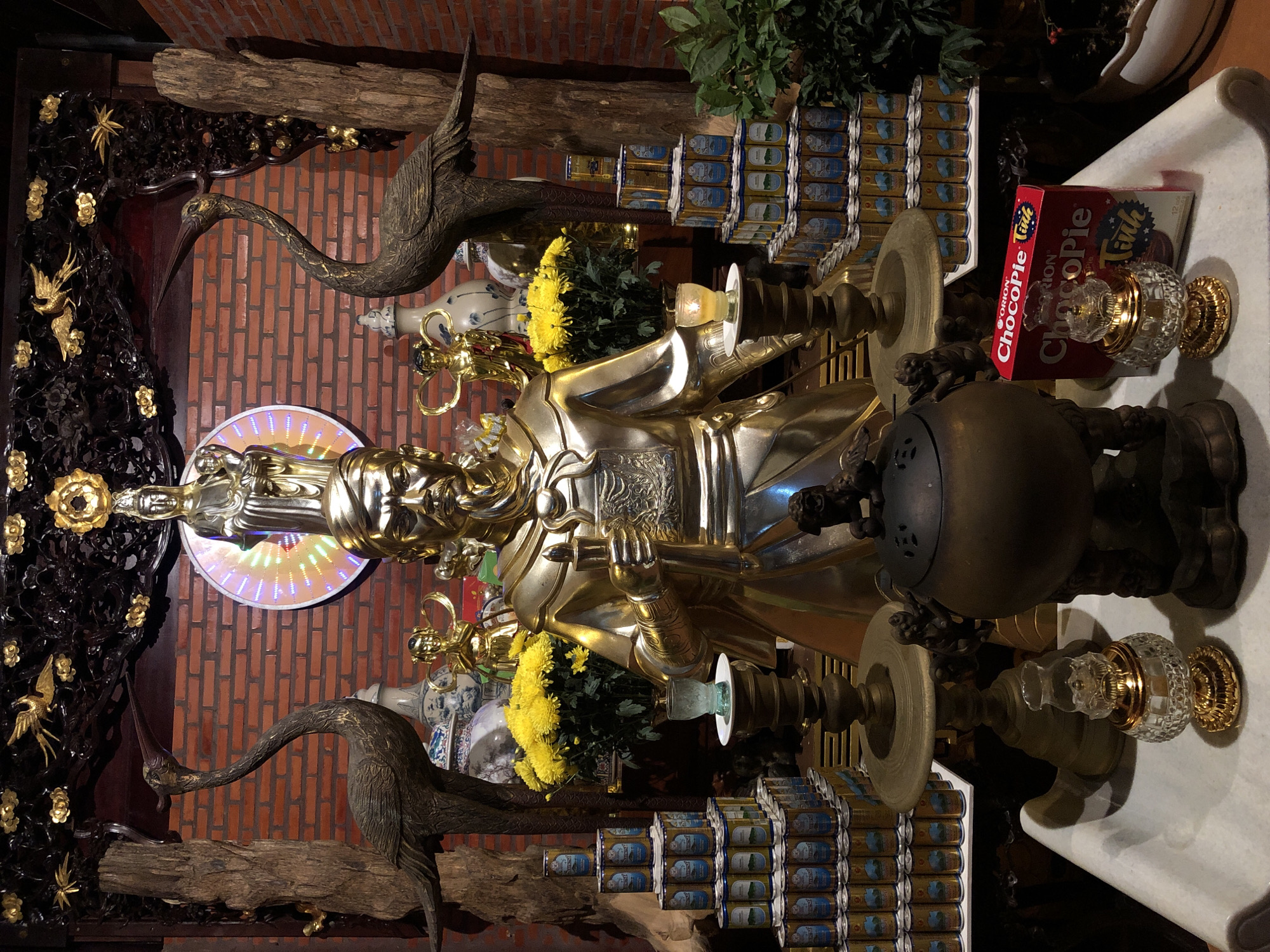 This is a photograph of an altar, likely in a temple or home, in Mũi Né, Vietnam. The altar is adorned with various devotional objects and decorations. Dominating the center is a large, golden statue of a deity or revered figure, seated and adorned in elaborate armor and headdress. To the left of the statue, a dark, intricately carved wooden panel features golden floral motifs and small bird figures. Two large, dark statues of cranes with long necks and slender beaks are positioned on either side of the central figure, one on the left and one on the right, seeming to guard the altar.
Behind the statue, a vase with blue and white patterns and a bouquet of vibrant yellow chrysanthemums are visible. The background consists of a brick wall, with a circular light fixture emitting a rainbow-like glow behind the deity. Stacked to the right of the altar are rows of what appear to be beverage cans, with a red box of "Orion ChocoPie" prominently displayed on top. The lighting suggests an indoor setting, possibly with artificial light sources illuminating the scene. The overall atmosphere is one of reverence and ornate display.