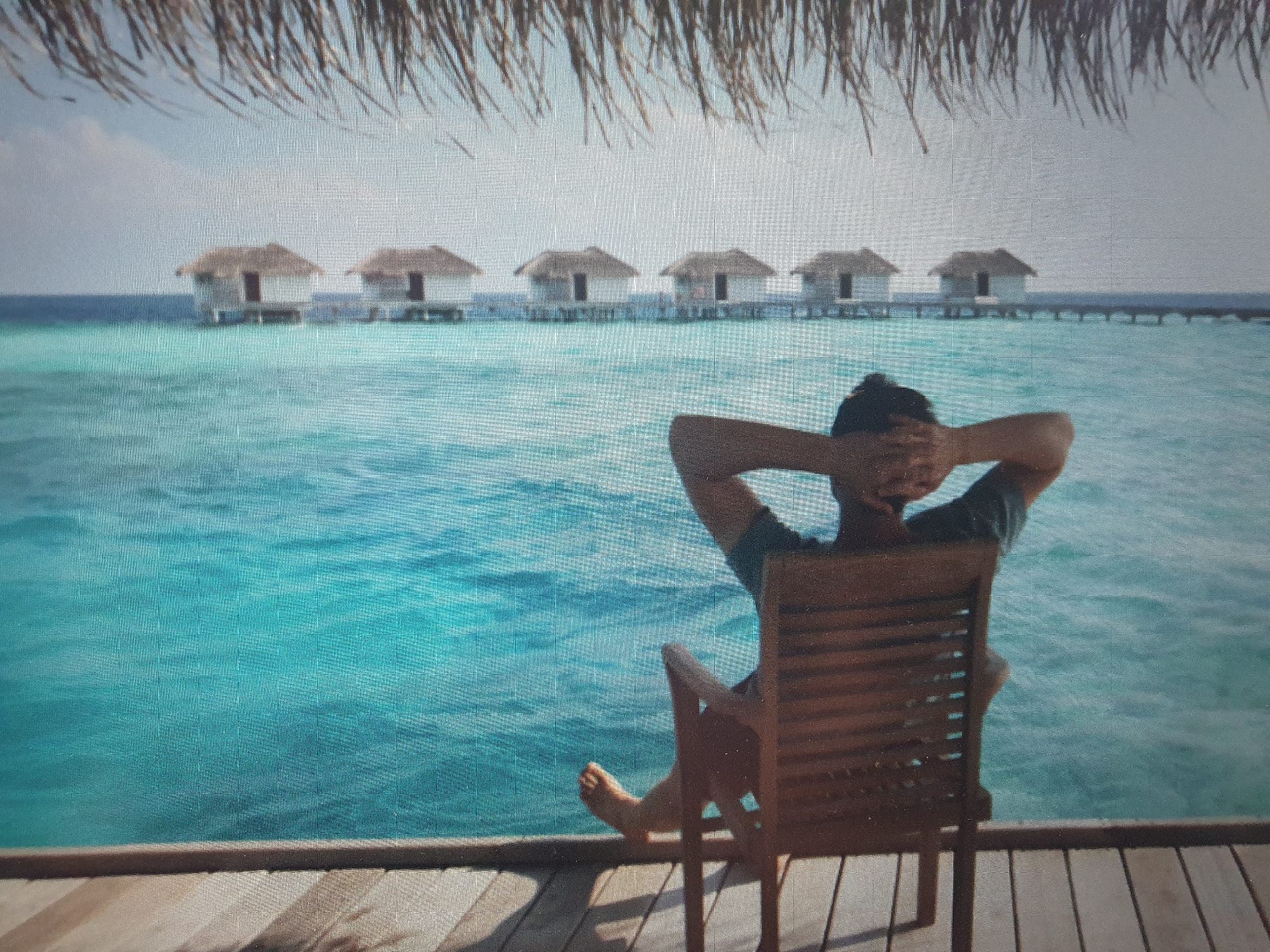 A person is sitting on a wooden chair on a wooden deck, looking out at the ocean. The person has their hands behind their head and is facing away from the camera. In the distance, there are several overwater bungalows. The water is a vibrant turquoise color, and the sky is clear with a few scattered clouds. The scene appears to be a tropical resort. There is no indication that this image is located in Tashkent, Uzbekistan.