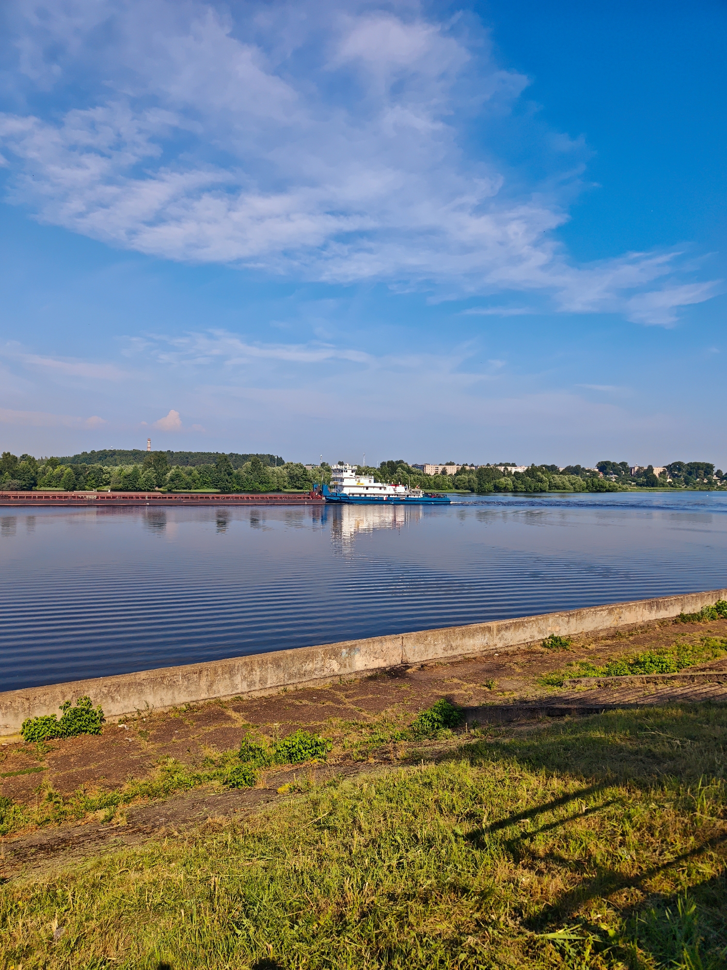 A cargo barge is seen on the Volga River in Kimry, Russia. The barge, painted white with blue trim, is being pushed by a tugboat. The river is wide and calm, with gentle ripples on its surface. The barge is loaded with what appear to be containers or other cargo. On the far bank of the river, a line of trees forms a green backdrop, with buildings and structures visible beyond the trees. The sky above is a bright blue with scattered white clouds. In the foreground, a concrete embankment runs along the edge of the river, with grass growing on its upper surface. The sun is shining, casting shadows on the grass.
